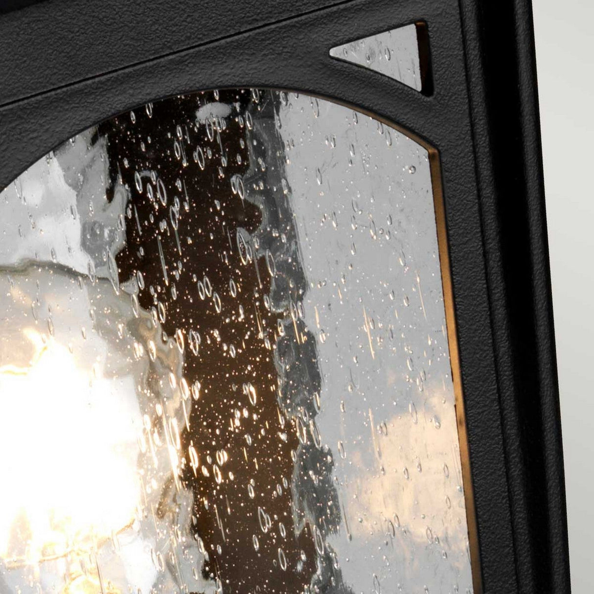 Close-up of a rain-splattered window with a blurred view of a bright light and trees in the background. The window frame enhances the vintage charm, contrasting with the glistening water droplets on the glass, reminiscent of an outdoor landscape illuminated by the Bedford 1 Light Medium Pedestal Lantern - Black.