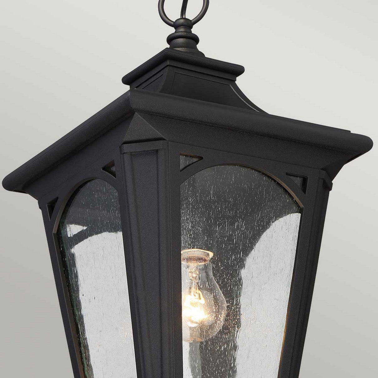 Close-up of the Bedford Outdoor 1 Light Small Chain Pendant - Black, a vintage-style lantern with an IP44 rating, suspended from above. It boasts a black metal frame and seedy glass panels, showcasing an illuminated light bulb inside. The background is a plain light gray.