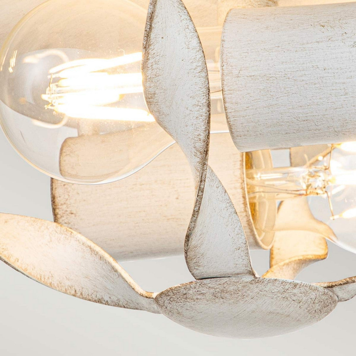 Close-up of the Bradbury 2 Light Flush - Antique White, showcasing two exposed bulbs framed by elegantly curved, leaf-like metal accents with a rustic, weathered finish. The bulbs emit a warm glow that enhances the vintage appeal and highlights the textured surface of the metallic elements.
