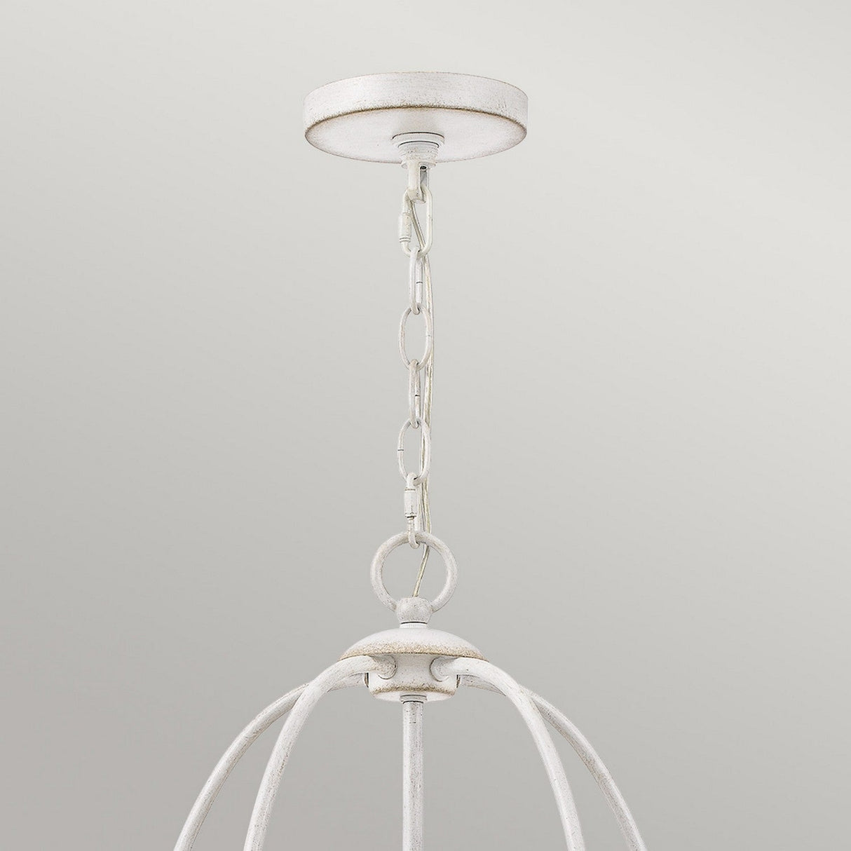 A close-up of an overhead light fixture exudes vintage charm with its rustic metal chain supporting an open, cage-like frame. The Bradbury 5 Light Chandelier - Antique White showcases a distressed finish, beautifully contrasting against a smooth, light gray background.