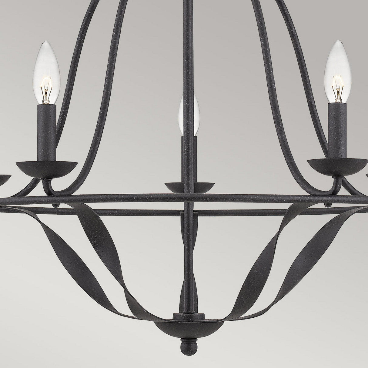 A close-up view of the Bradbury 5 Light Chandelier in Grey Ash reveals its contemporary flair with three candle-like bulbs. The minimalist design showcases curved metal details reminiscent of the Grey Ash finish, all set against a light gray background.