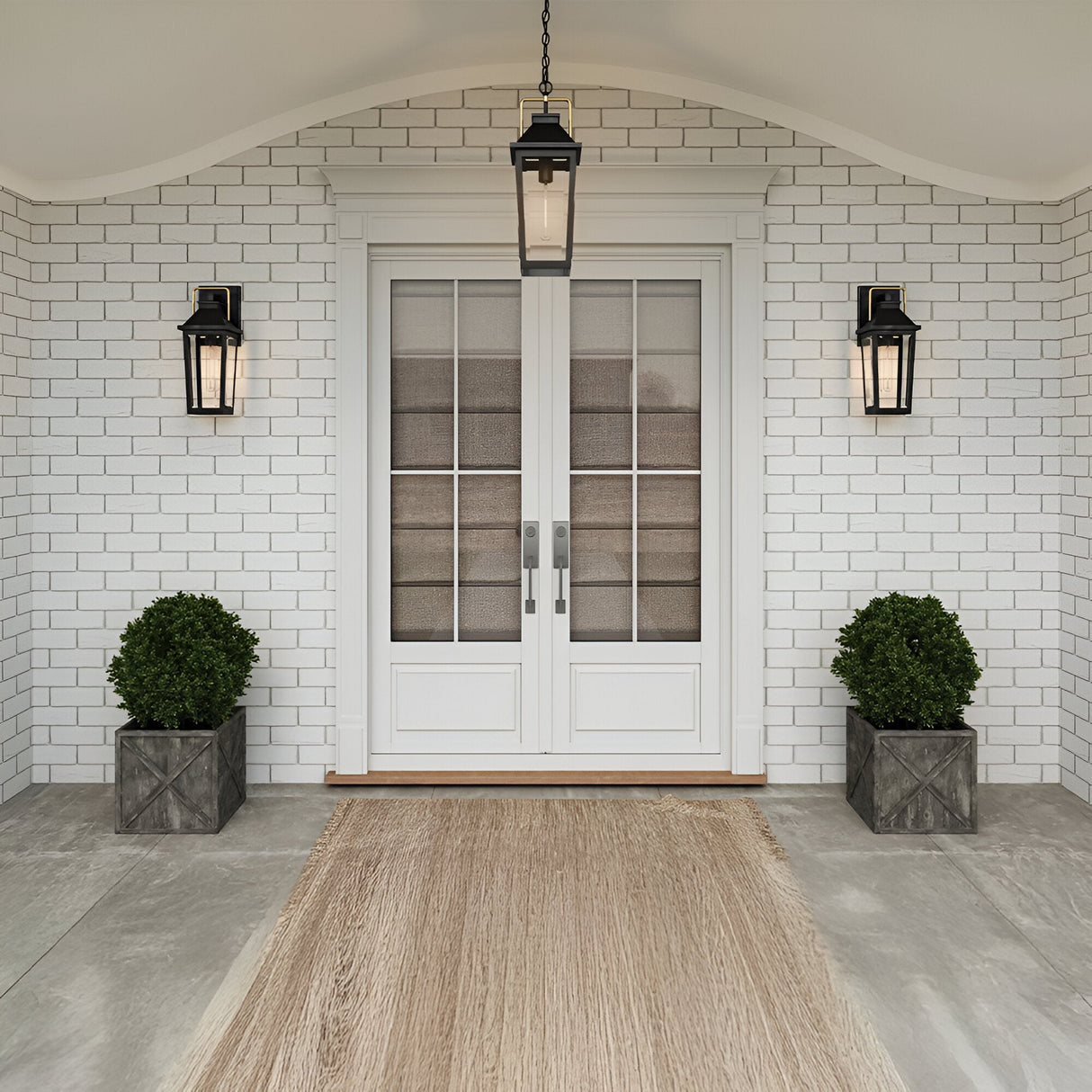 White brick entryway with double glass doors, two Buckley 1 Light Coastal Medium Outdoor Wall Lights Lanterns in black on each side, and two potted round shrubs in square planters. A beige doormat sits on the gray concrete porch.