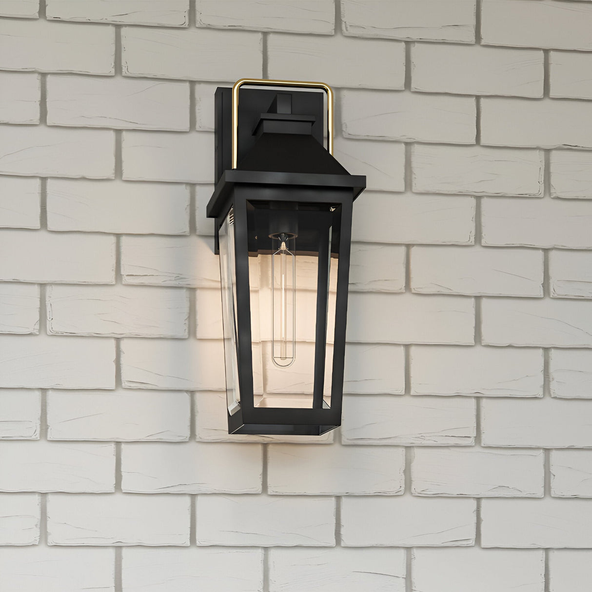 The Buckley 1 Light Coastal Medium Outdoor Wall Lights Lantern in black, featuring clear glass panels and gold detail, is mounted on a light gray brick wall. This weatherproof fixture showcases a modern, geometric design with a visible bulb.