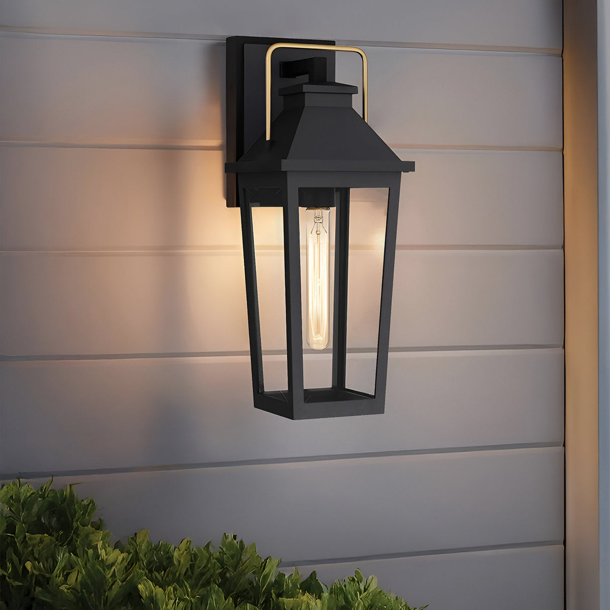 The Buckley 1 Light Coastal Medium Outdoor Wall Lights Lantern in black is mounted on grey siding, illuminating the area. This weatherproof fixture brightens the space, with greenery visible below.