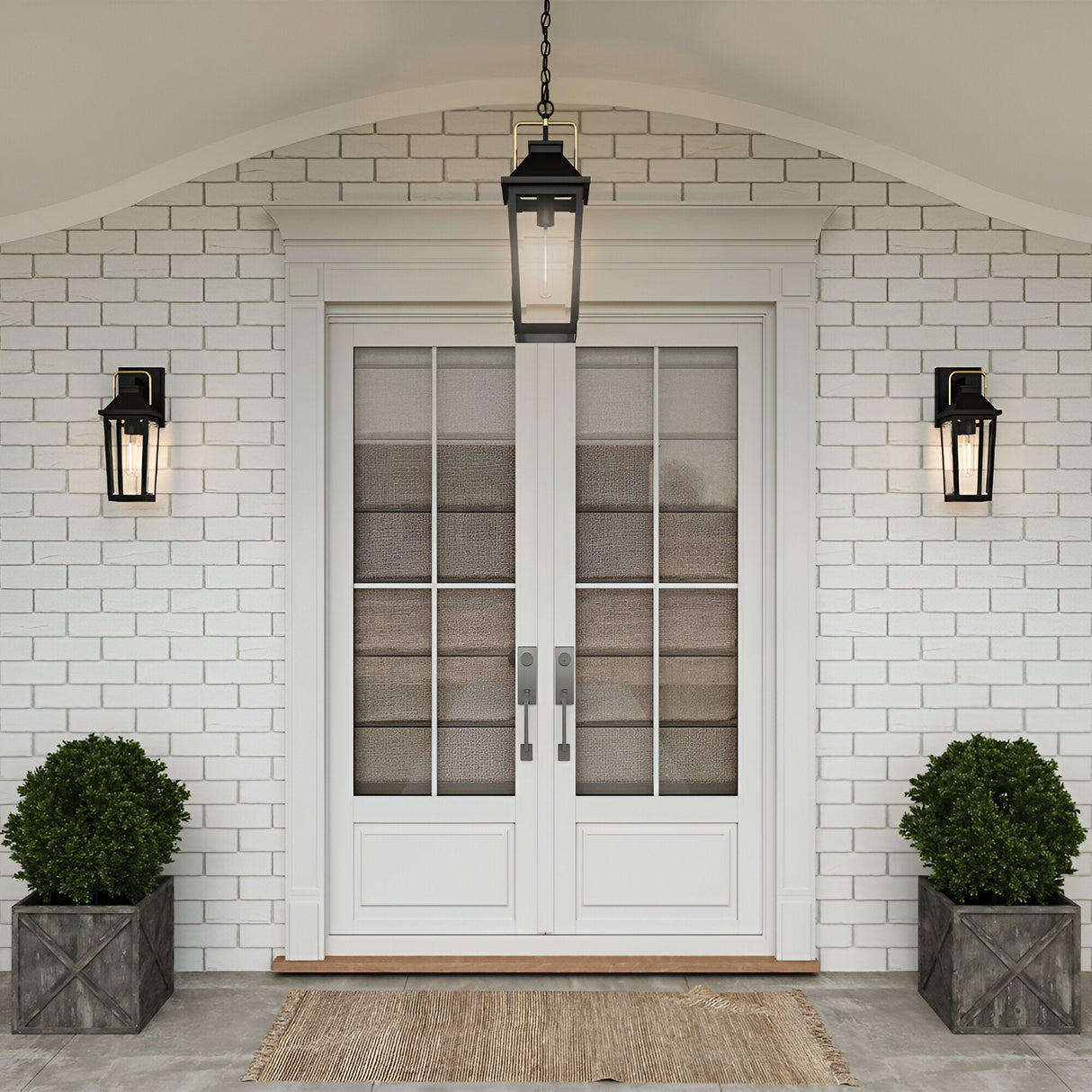 White double front doors with glass panels and silver handles are accented by a Buckley 1 Light Coastal Small Outdoor Wall Lights Lantern in black and potted shrubs in square planters. A brown doormat sits on the brick porch below a stylish hanging lantern above.