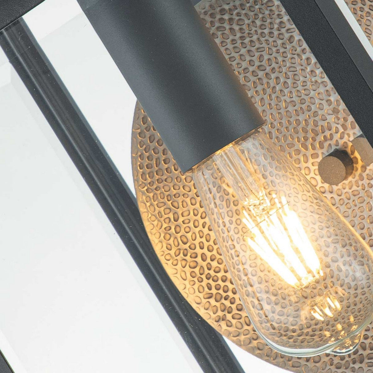 A close-up of an illuminated Edison-style light bulb with visible filaments is set against a textured, circular metal backplate. The wall lantern features a black cylindrical holder and is partially enclosed by bevelled glass panels in a graphite finish, capturing the charm of the Byron Outdoor 1 Light Large Wall Lantern - Graphite With Grey Reflector.