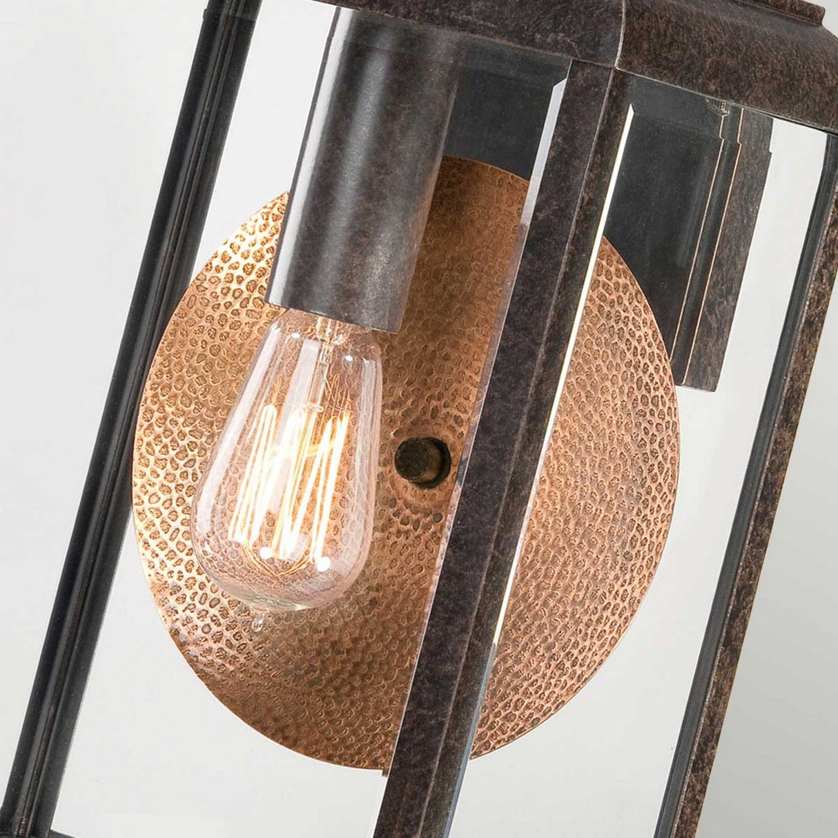Close-up of the Byron Outdoor 1 Light Large Wall Lantern in Imperial Bronze, showcasing a textured bronze circular backplate and an exposed Edison bulb. Its rectangular glass and metal frame, complemented by a copper reflector, emits a warm glow.