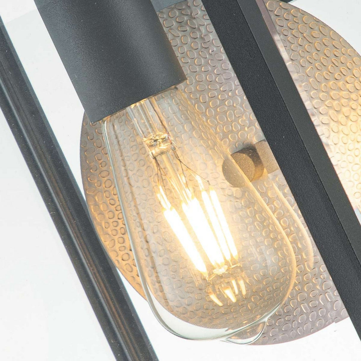 A close-up of a lit vintage-style light bulb with visible filaments is set against a textured metallic background, enclosed in a black metal frame that evokes the sleek design of the Byron Outdoor 1 Light Medium Wall Lantern in Graphite with Grey Reflector, creating a modern industrial look.