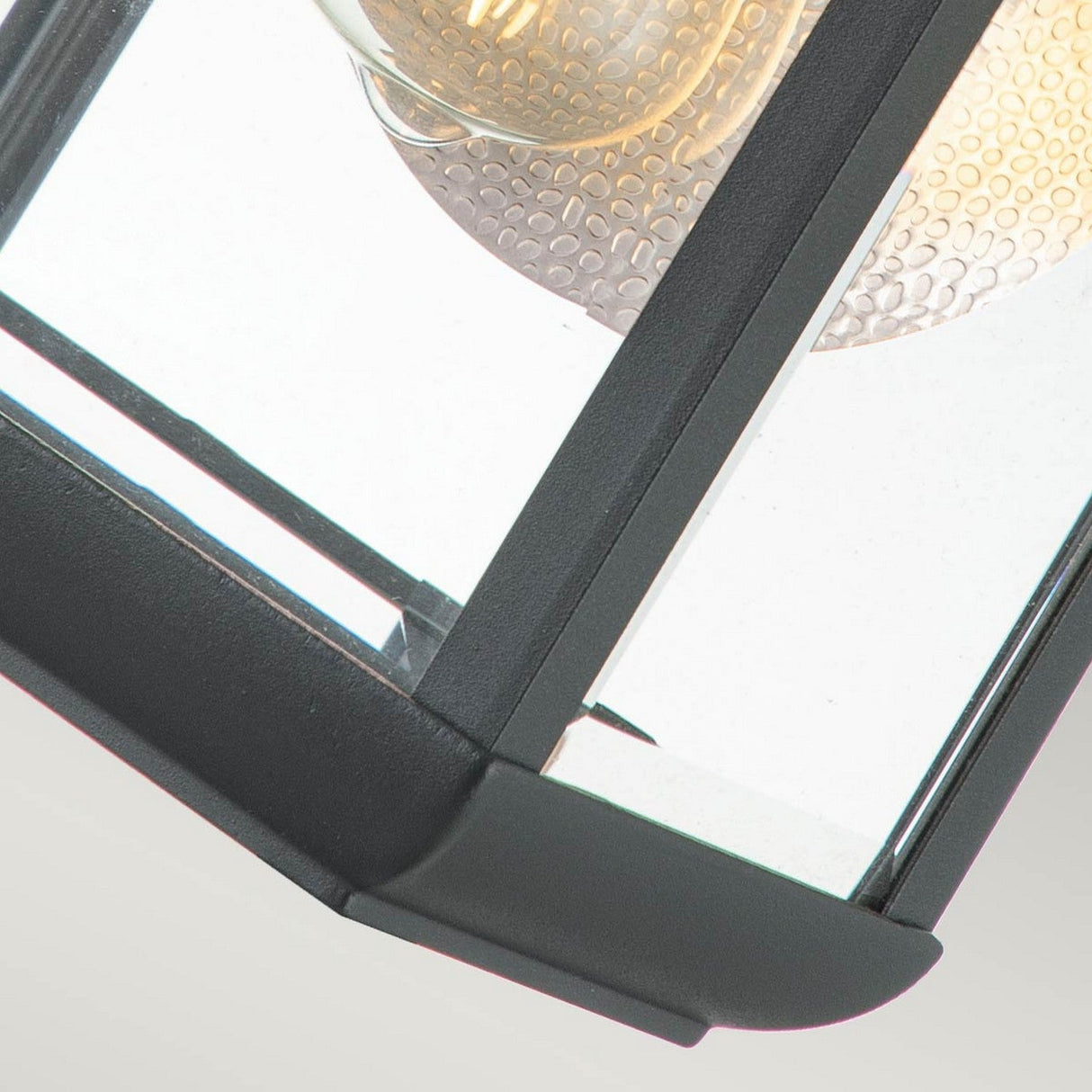 Close-up of the Byron Outdoor 1 Light Medium Wall Lantern in Graphite, featuring a black metal frame and clear glass panels. Inside, a textured, spherical light bulb emits a warm glow. The image highlights the corner of this IP44-rated lantern with its grey reflector against a light background, emphasizing its modern elegance in outdoor lighting.