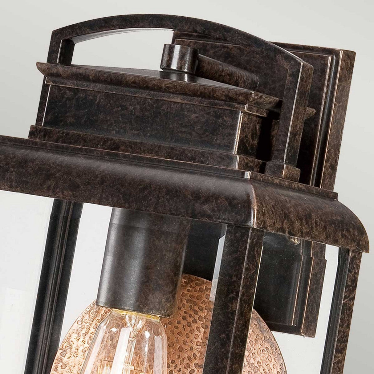 Close-up of the Byron Outdoor 1 Light Medium Wall Lantern in Imperial Bronze. The lantern's design features clear glass panes and a vintage-style exposed bulb, emanating rustic elegance. A copper reflector enhances the warm glow, set against a softly blurred background that completes the tranquil scene.