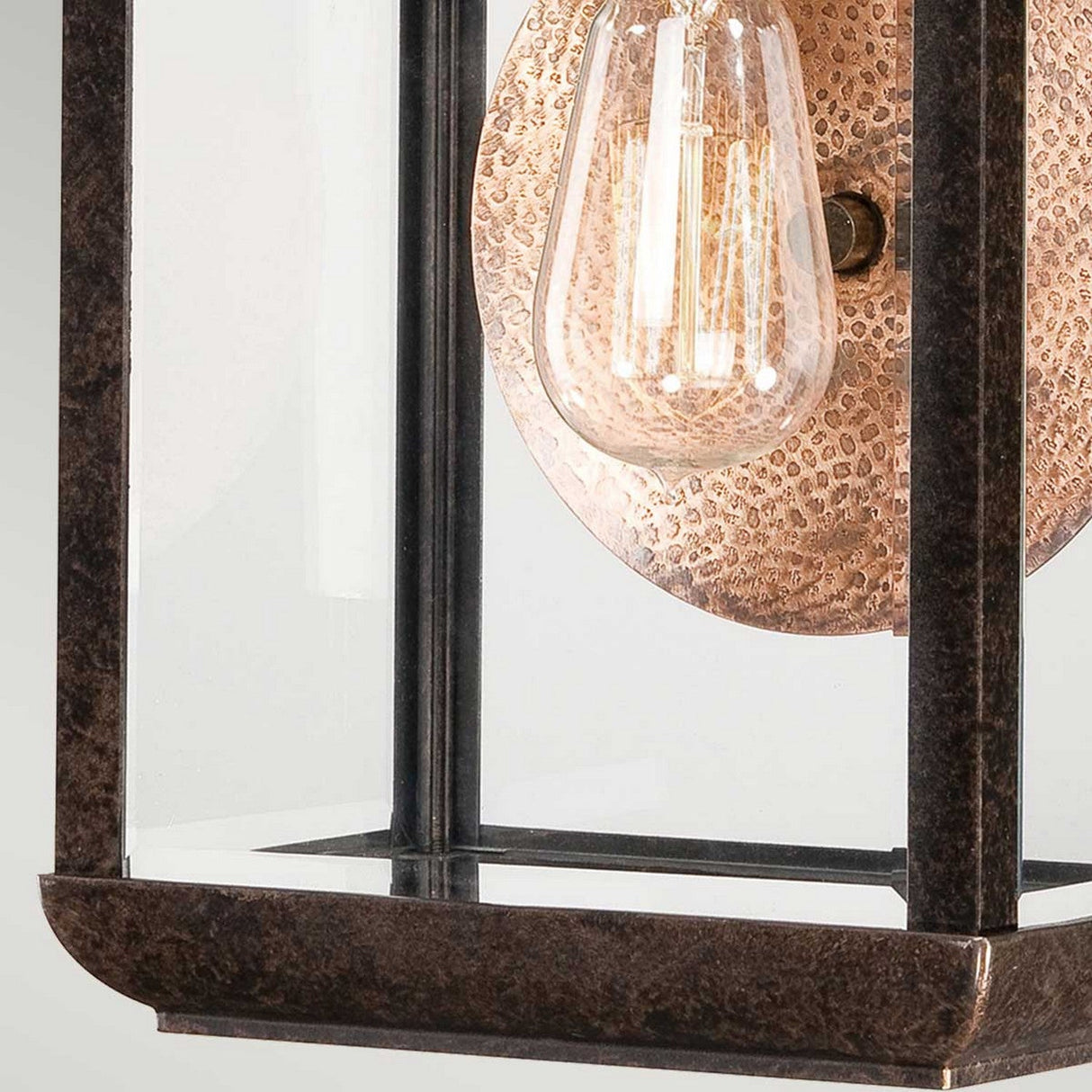 Close-up of the Byron Outdoor 1 Light Medium Wall Lantern, showcasing a vintage-style light fixture with a clear glass enclosure, an exposed filament bulb, and a textured backplate in Imperial Bronze with a copper reflector finish, set against a neutral background.