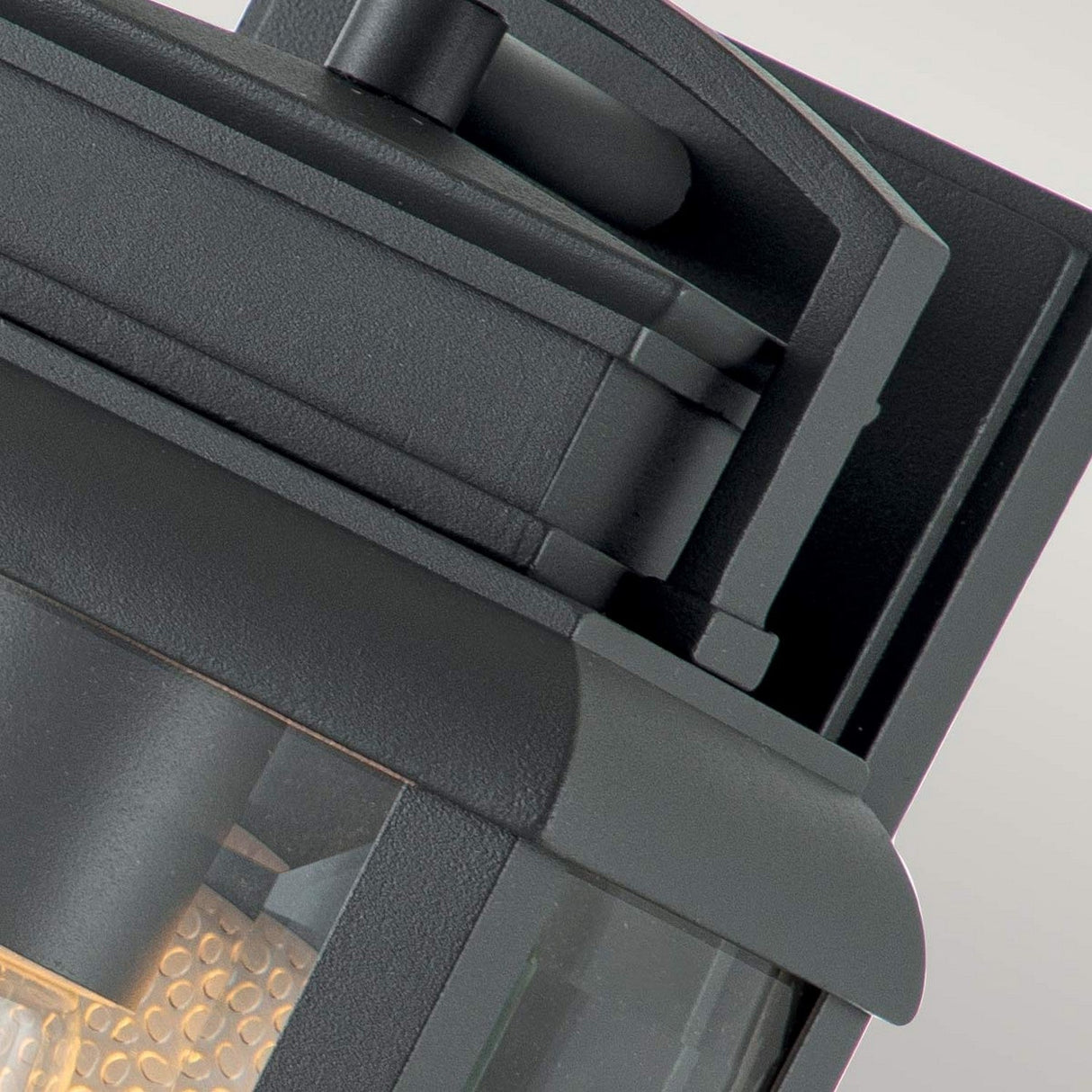 Close-up of the Byron Outdoor 1 Light Small Wall Lantern, showcasing a graphite finish with a grey reflector and a rectangular design. The fixture's transparent glass panels and textured appearance emit a warm glow. Its compact and sleek structure is mounted on a neutral-colored wall and is IP44-rated for durability.