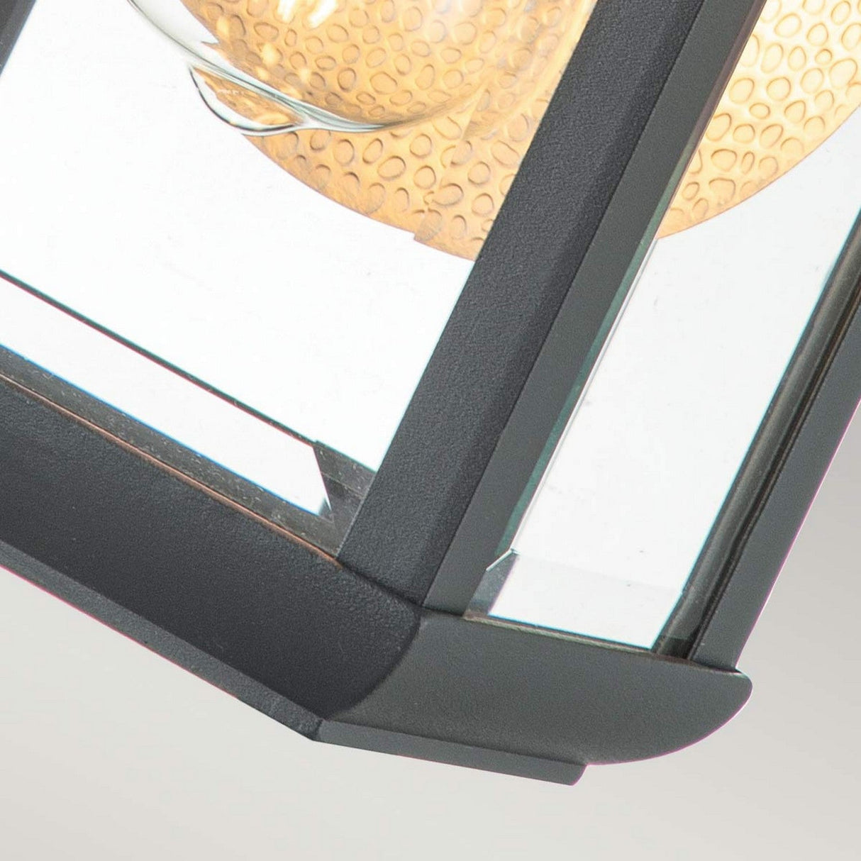 Close-up of the Byron Outdoor 1 Light Small Wall Lantern, featuring a sleek black metal frame and clear glass panels in a graphite finish. The interior boasts a textured beige sphere as the light source, providing an eye-catching contrast to the modern design of this IP44-rated lantern.