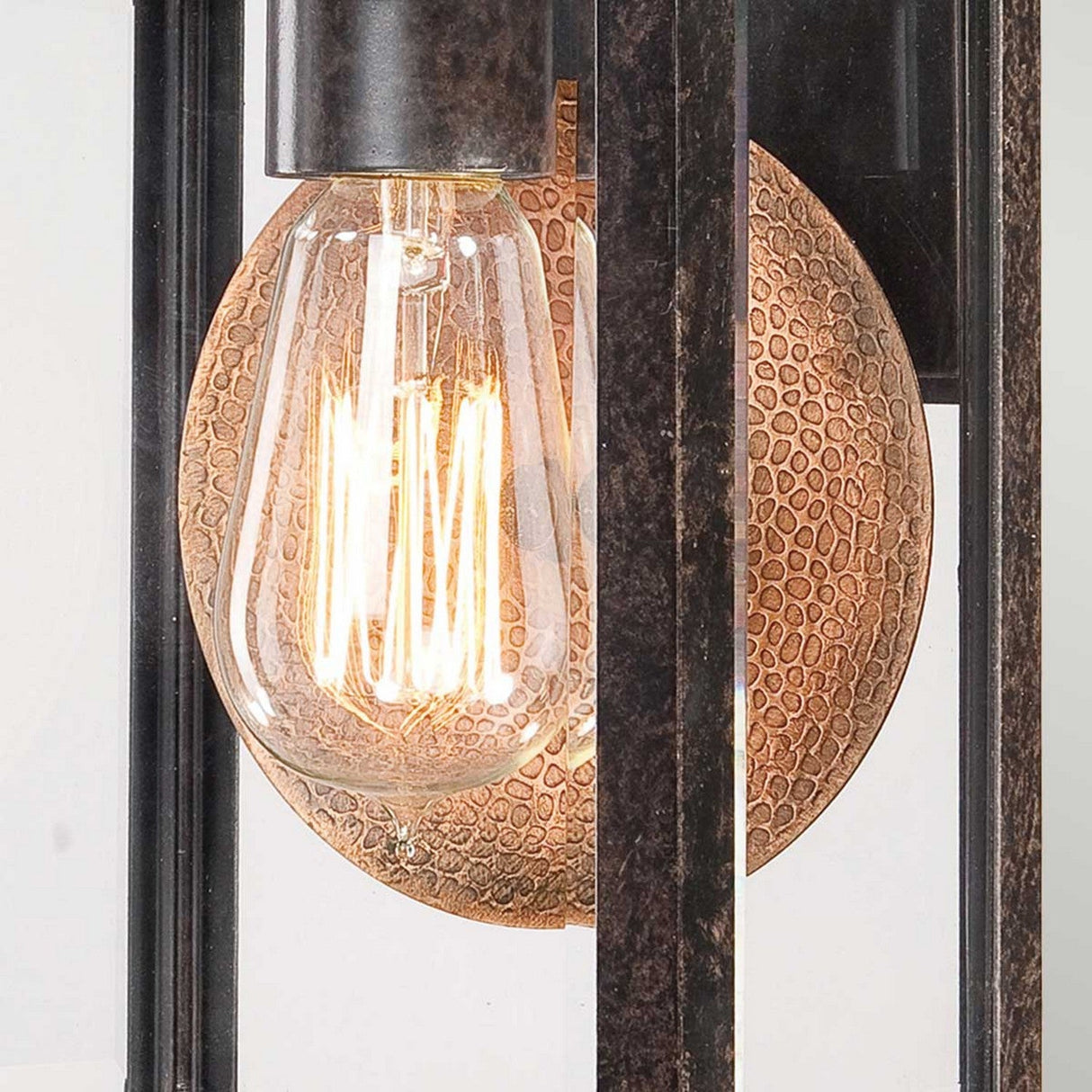 The Byron Outdoor 1 Light Small Wall Lantern features a close-up of an Edison-style light bulb with visible filaments encased in a glass cover. The bulb is mounted on a textured copper reflector and framed by the lantern's dark metal structure, offering timeless elegance with its Imperial Bronze finish.