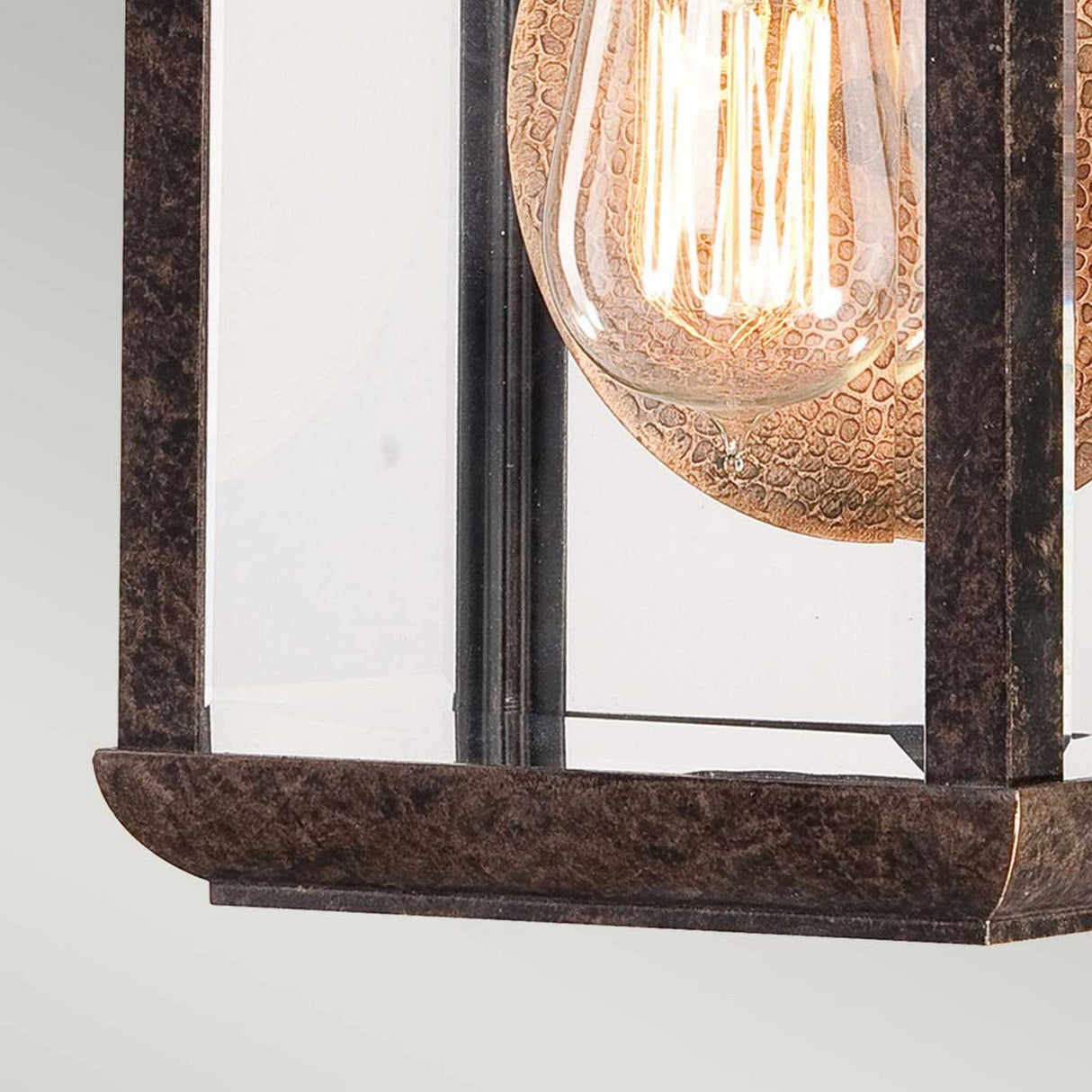 Close-up of the Byron Outdoor 1 Light Small Wall Lantern featuring a vintage-style design with a visible filament bulb. The fixture, encased in glass and metal with an Imperial Bronze finish and copper reflector, showcases intricate design details that evoke timeless elegance.