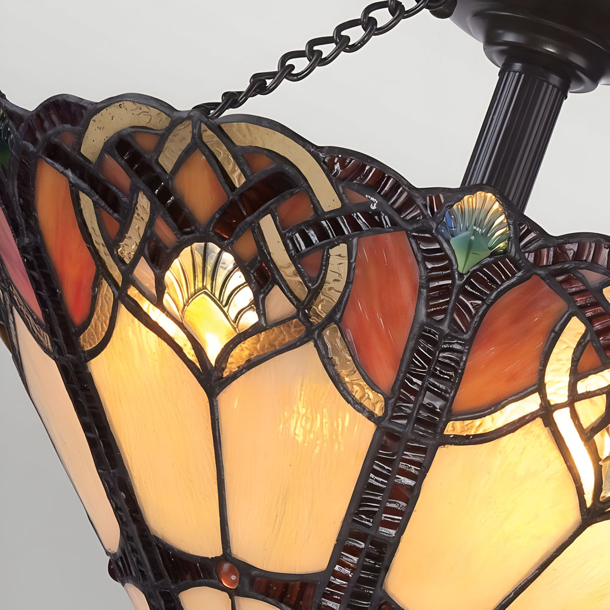 Close-up of the Cambridge 2 Light Large Tiffany Semi-Flush in Vintage Bronze, featuring a stained glass shade with intricate patterns in orange, yellow, and green hues, accentuated by dark outlines. The design showcases floral and geometric shapes beautifully illuminated by a soft, warm light from behind.