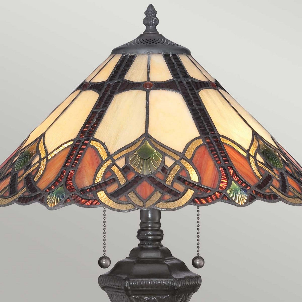 The Cambridge 2 Light Table Lamp in vintage bronze highlights intricate patterns in warm tones of cream, orange, and green on its decorative stained glass shade. It includes pull chains and is elegantly set against a plain background.