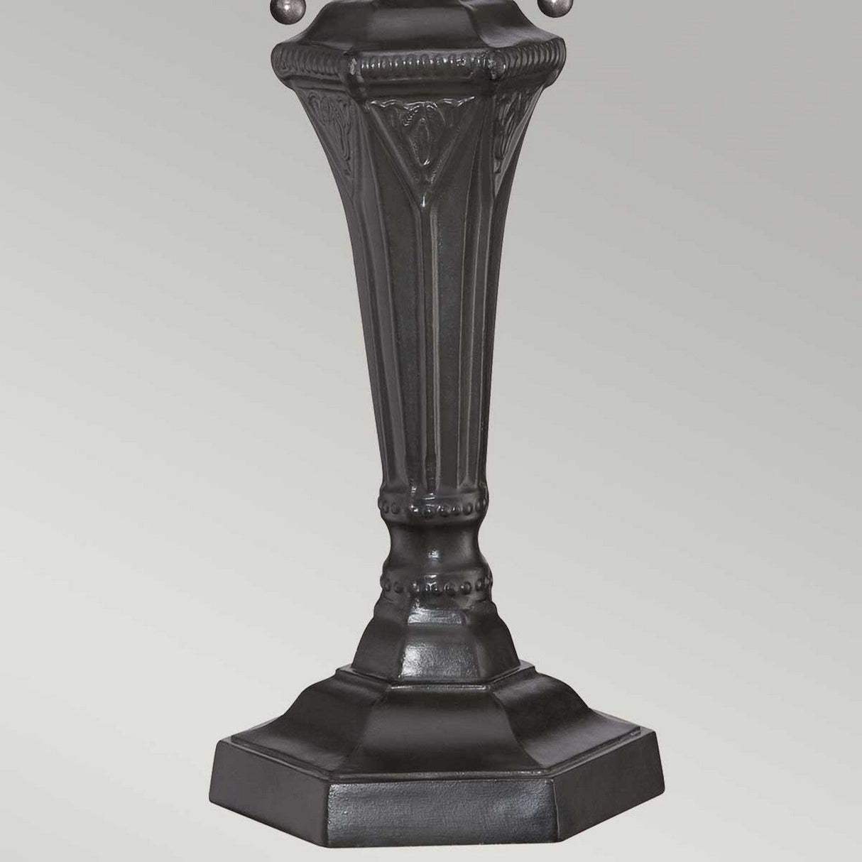A close-up of the Cambridge 2 Light Table Lamp reveals its dark, ornate base adorned with intricate carvings. This vintage bronze base boasts a hexagonal shape and a robust, antique design, highlighted by detailed vertical grooves and decorative elements. The stained glass shade casts vibrant colors against the plain gray background.