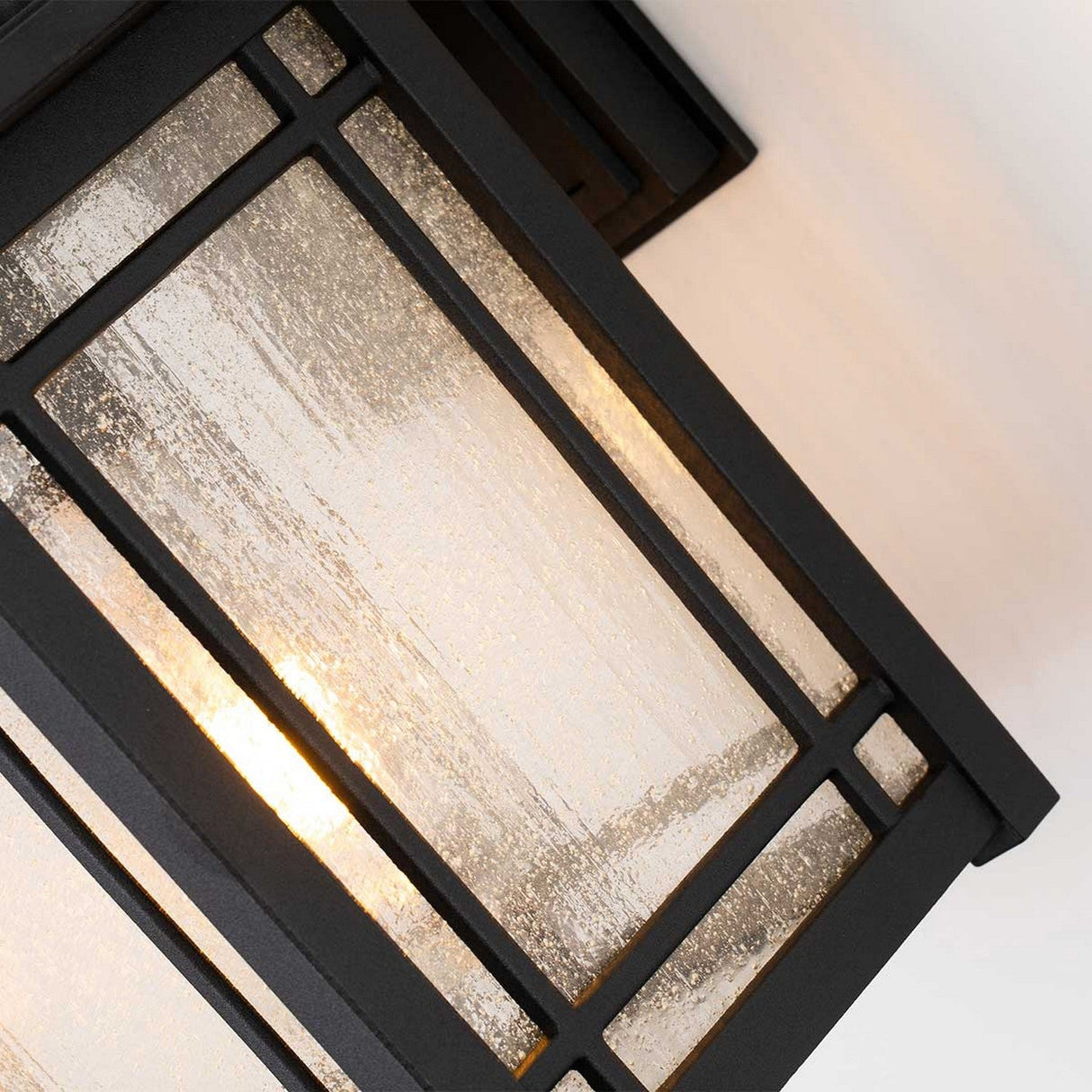 Close-up of the Cedar Point Outdoor 1 Light Medium Wall Lantern - Black, showcasing its black metal frame and seedy glass panels, illuminated from within. The light emits a warm glow, accentuating the textured patterns on the glass, making it ideal for outdoor lighting.