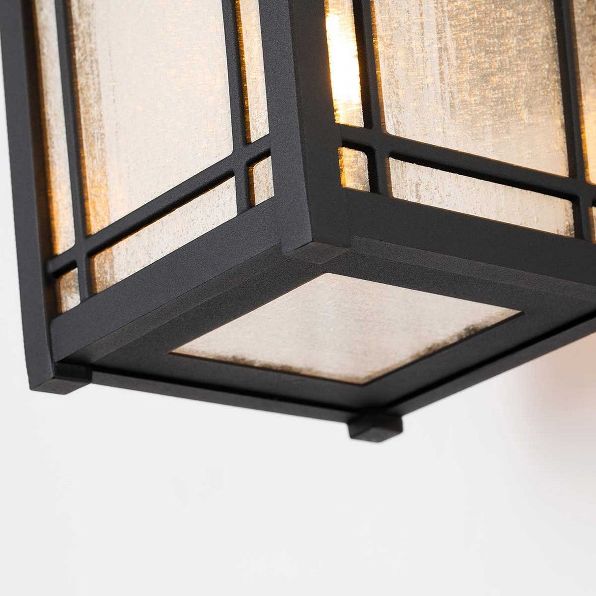 Close-up of a contemporary, geometric black metal light fixture featuring seedy glass panels, gently lit from the inside. It projects a warm glow against a plain white backdrop, echoing the design of the Cedar Point Outdoor 1 Light Medium Wall Lantern - Black in its elegant outdoor lighting style.