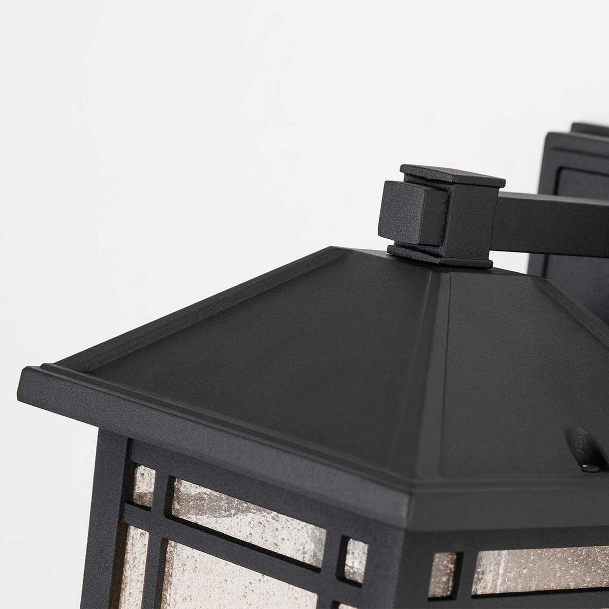 Close-up of the Cedar Point Outdoor 1 Light Small Wall Lantern - Black, highlighting its black rectangular metal frame and seedy glass panels. It features a pyramid-shaped top and is mounted on a wall bracket, providing rustic outdoor lighting. The background is plain white.