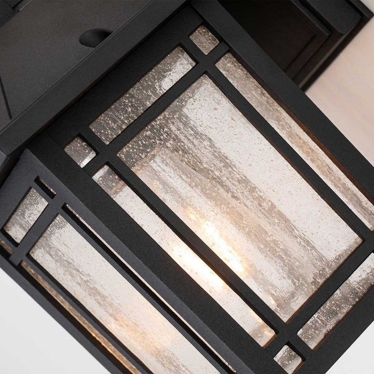 Detailed view of the Cedar Point Outdoor 1 Light Small Wall Lantern - Black featuring a rectangular design. The black metal frame encloses seedy glass panels, allowing warm light to shine through and create an inviting ambiance perfect for rustic outdoor lighting.