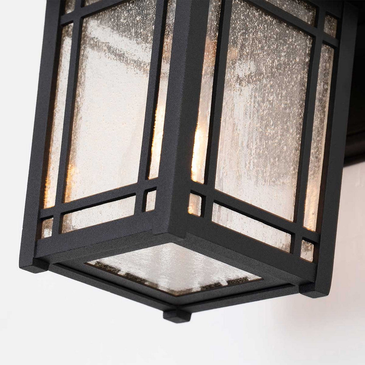 Close-up of the Cedar Point Outdoor 1 Light Small Wall Lantern - Black, featuring a geometric design and mounted on a wall. The seedy glass panels exhibit a textured, speckled appearance, while the rustic outdoor lighting emits a warm glow.
