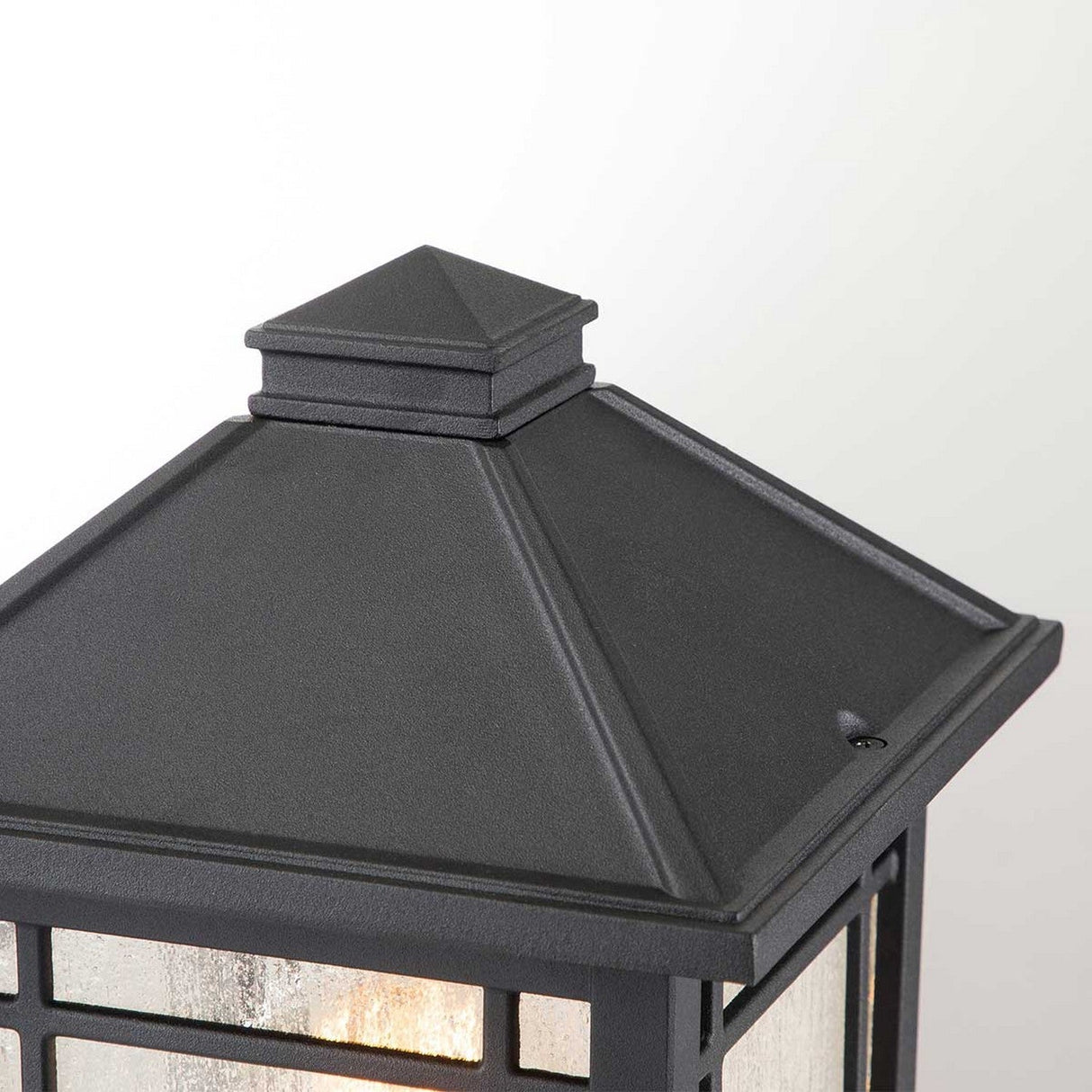The Cedar Point 1 Light Pedestal - Black boasts vintage charm with its pyramid design and textured glass panels. Mounted against a light-colored backdrop, it casts a warm glow that beautifully enhances any outdoor pathway.