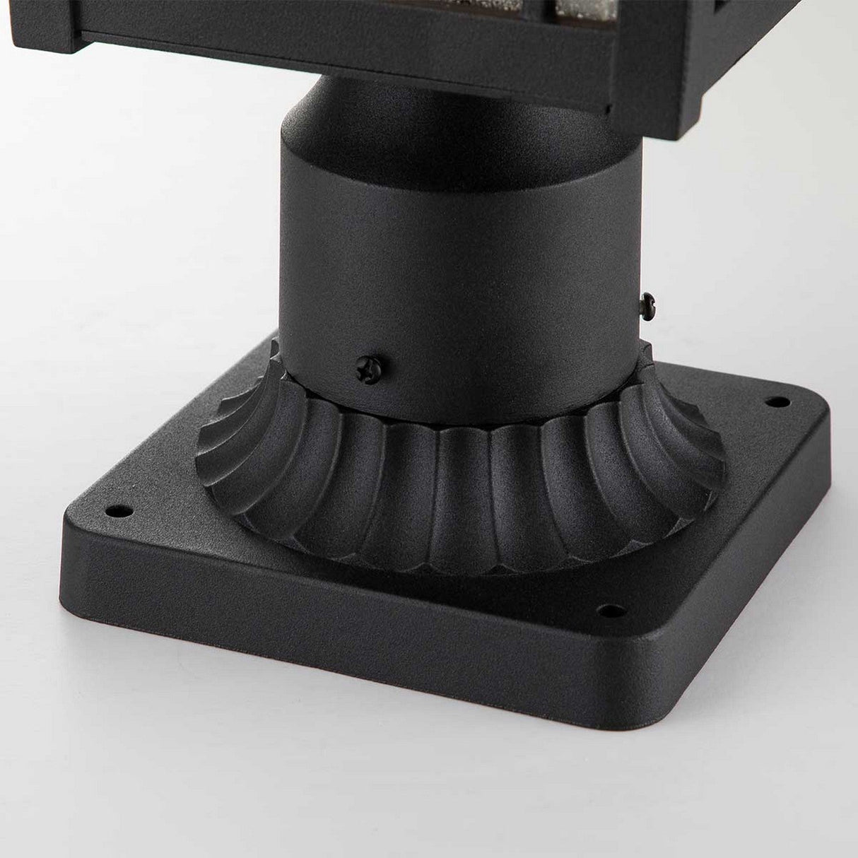 A close-up of the Cedar Point 1 Light Pedestal - Black showcases a decorative grooved design on its black metal base, exuding vintage character. Mounted on a flat surface, this square pedestal offers a minimalist and industrial style perfect for outdoor pathways.