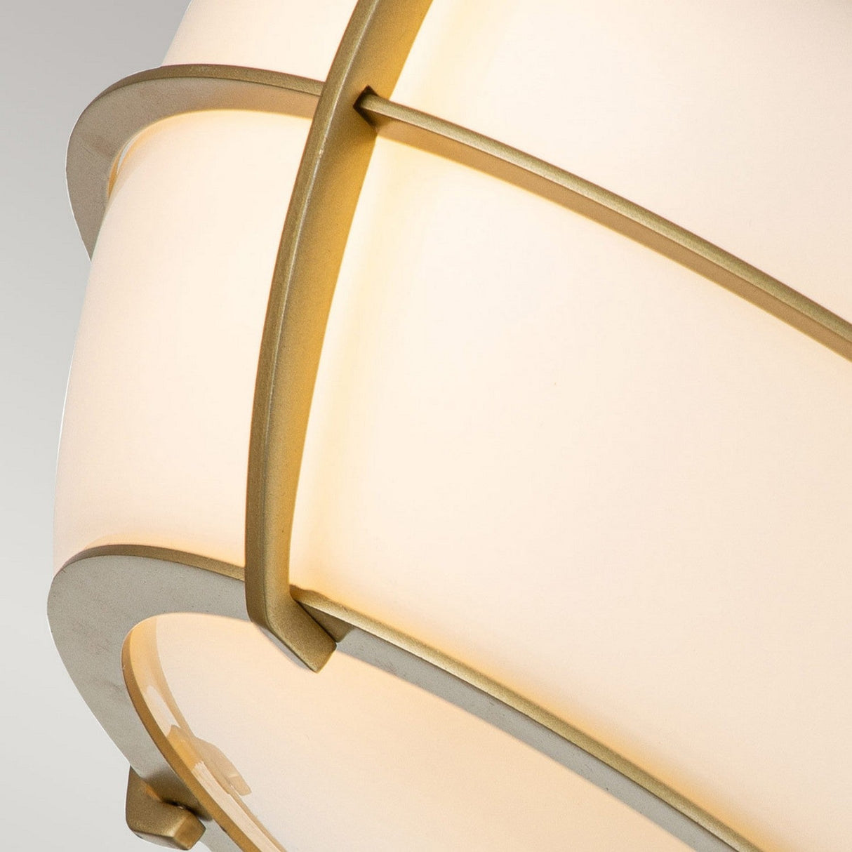 Close-up of the Chance 2 Light Flush - Natural Brass ceiling fixture, showcasing a spherical white shade with sleek natural brass bands arranged in a geometric pattern. The light casts a warm glow, accentuating the elegant design against a neutral background.