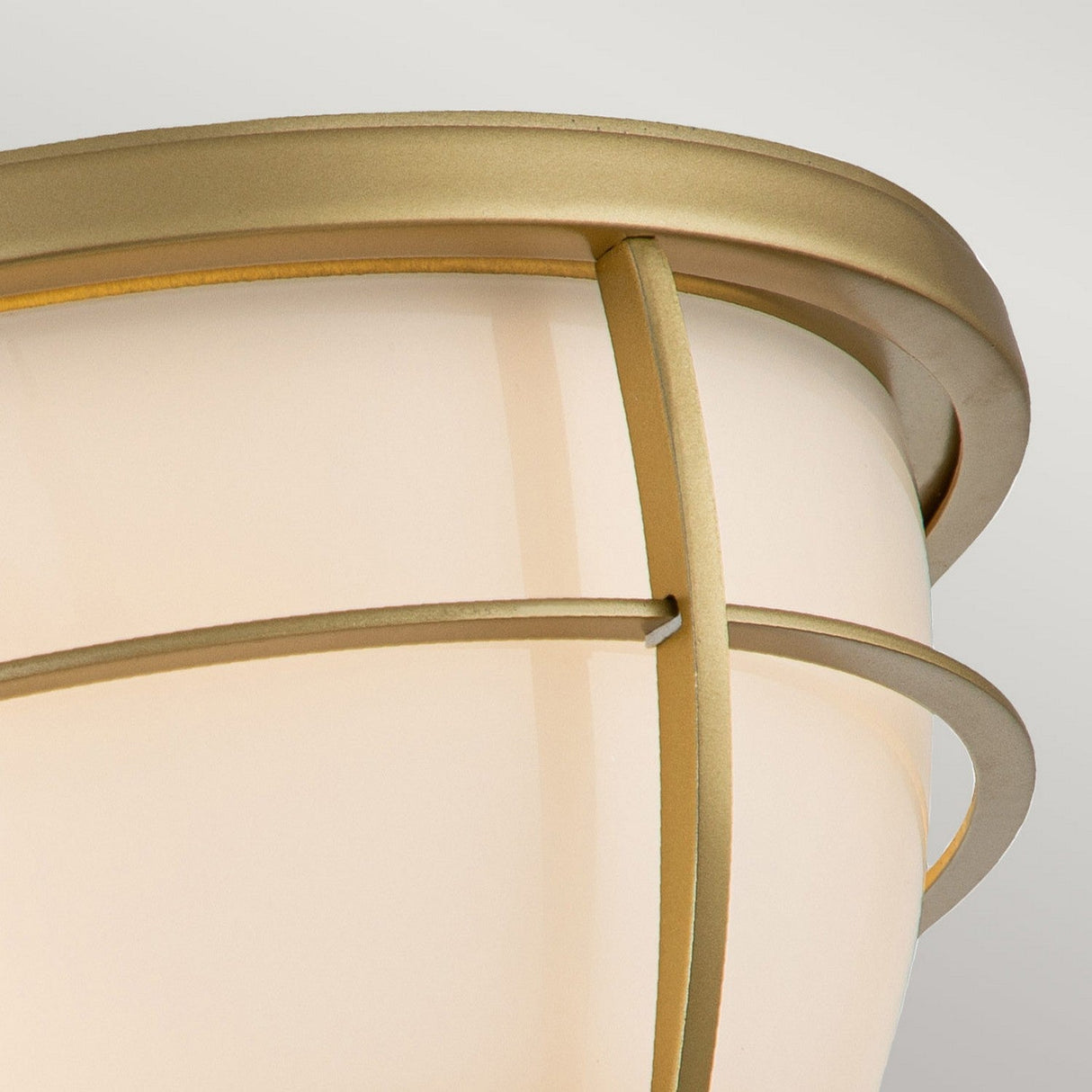 A close-up view of the Chance 2 Light Flush ceiling fixture in natural brass, showcasing frosted glass and intersecting metal bands that create geometric patterns. Its IP44 rating guarantees durability, and the diffused light provides a warm and elegant glow.