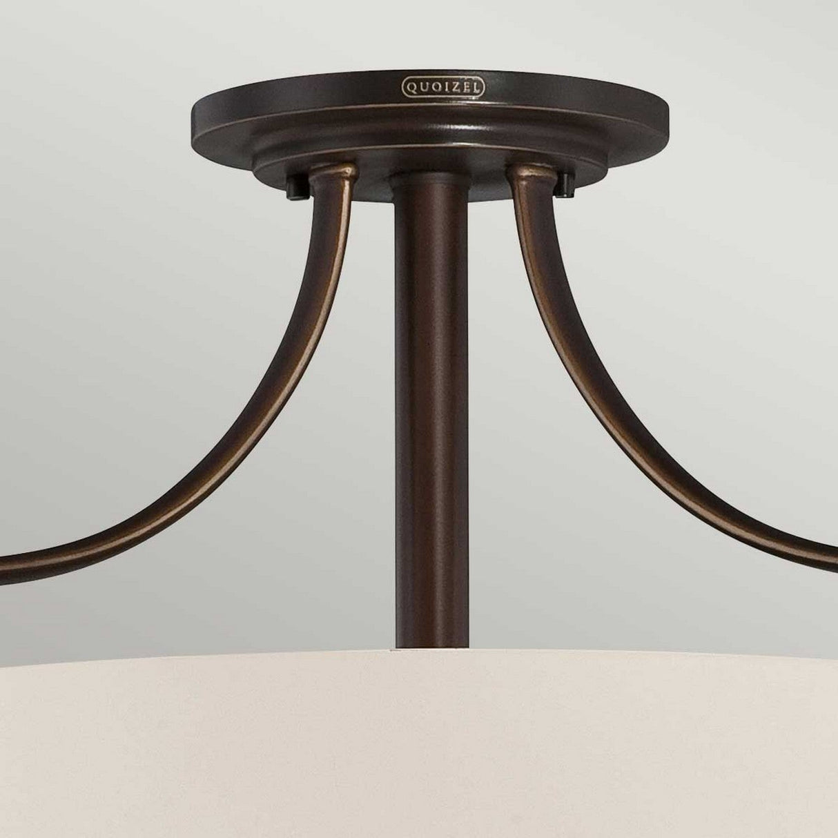 A close-up view of the Chantilly 3 Light Semi-Flush fixture showcases its Palladian Bronze finish and elegantly curved metal supports. This Quoizel piece features an opal-etched glass diffuser, highlighting its simple, modern design with the brand name displayed on the round base.