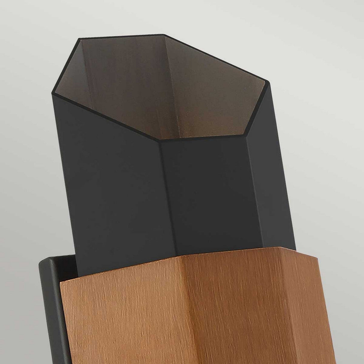 Close-up of a contemporary vase with a geometric pentagon shape. The base is crafted from wood, contrasting with the matte black metal top section that evokes the Chasm Outdoor LED Medium Wall Lantern. The design showcases clean lines and a minimalist aesthetic, set against a neutral backdrop.
