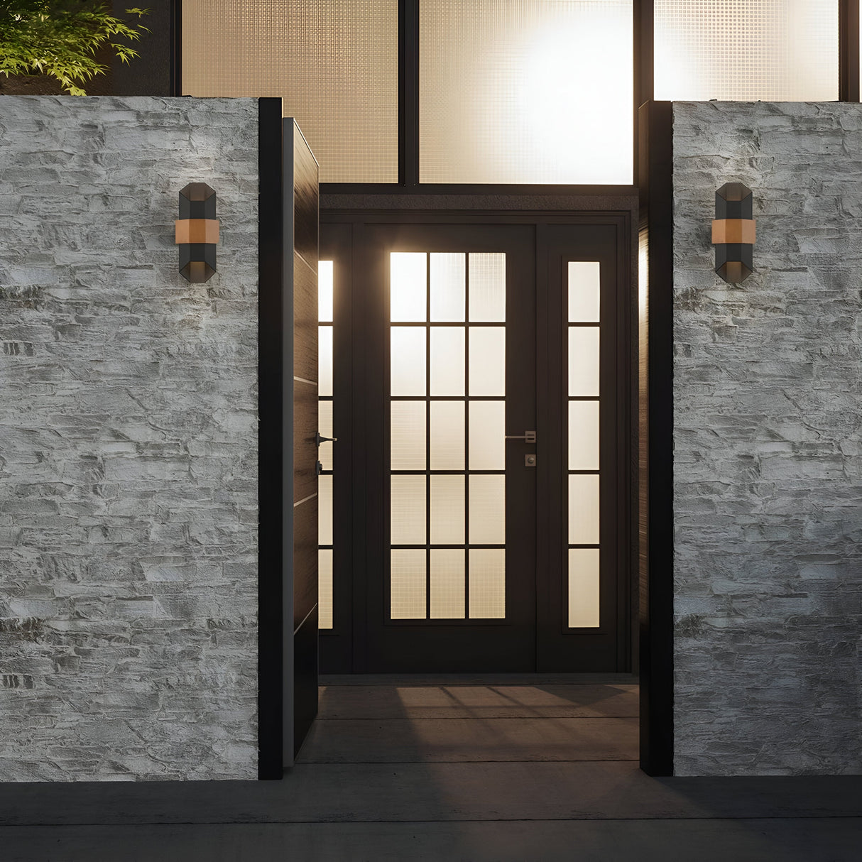 A contemporary entrance showcases a black-framed glass door adorned with geometric designs, nestled between two stone walls. The area is illuminated by Chasm Outdoor LED Medium Wall Lanterns in Matte Black, and greenery can be seen over the left wall. Natural light streams through the glass panels.