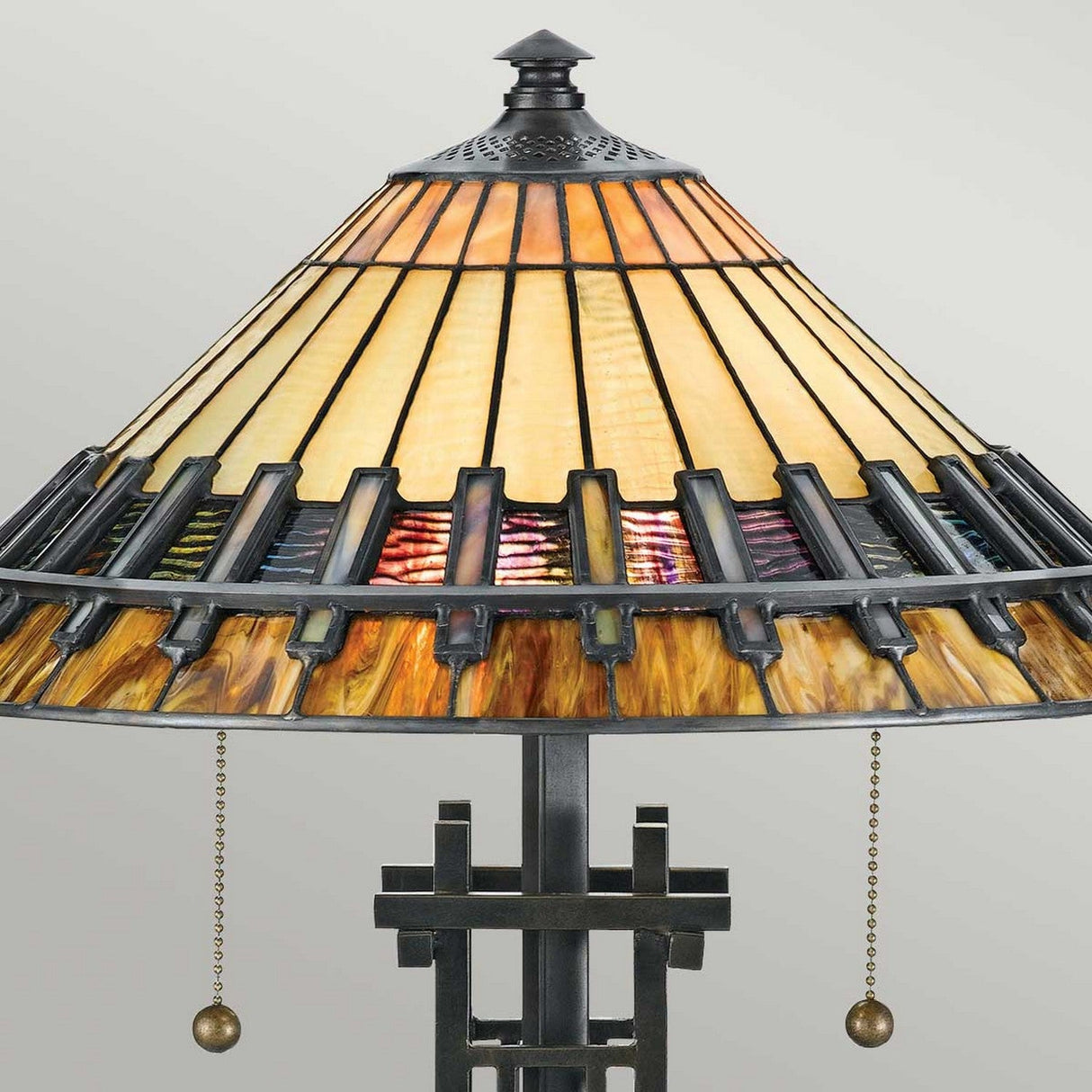 The Chastain 2 Light Tiffany Table Lamp in Vintage Bronze exudes Arts and Crafts elegance with its geometric design in warm earth tones. Its stained glass shade, framed in black metal and featuring two pull chains, sits gracefully against a plain gray background, highlighting its vintage charm.