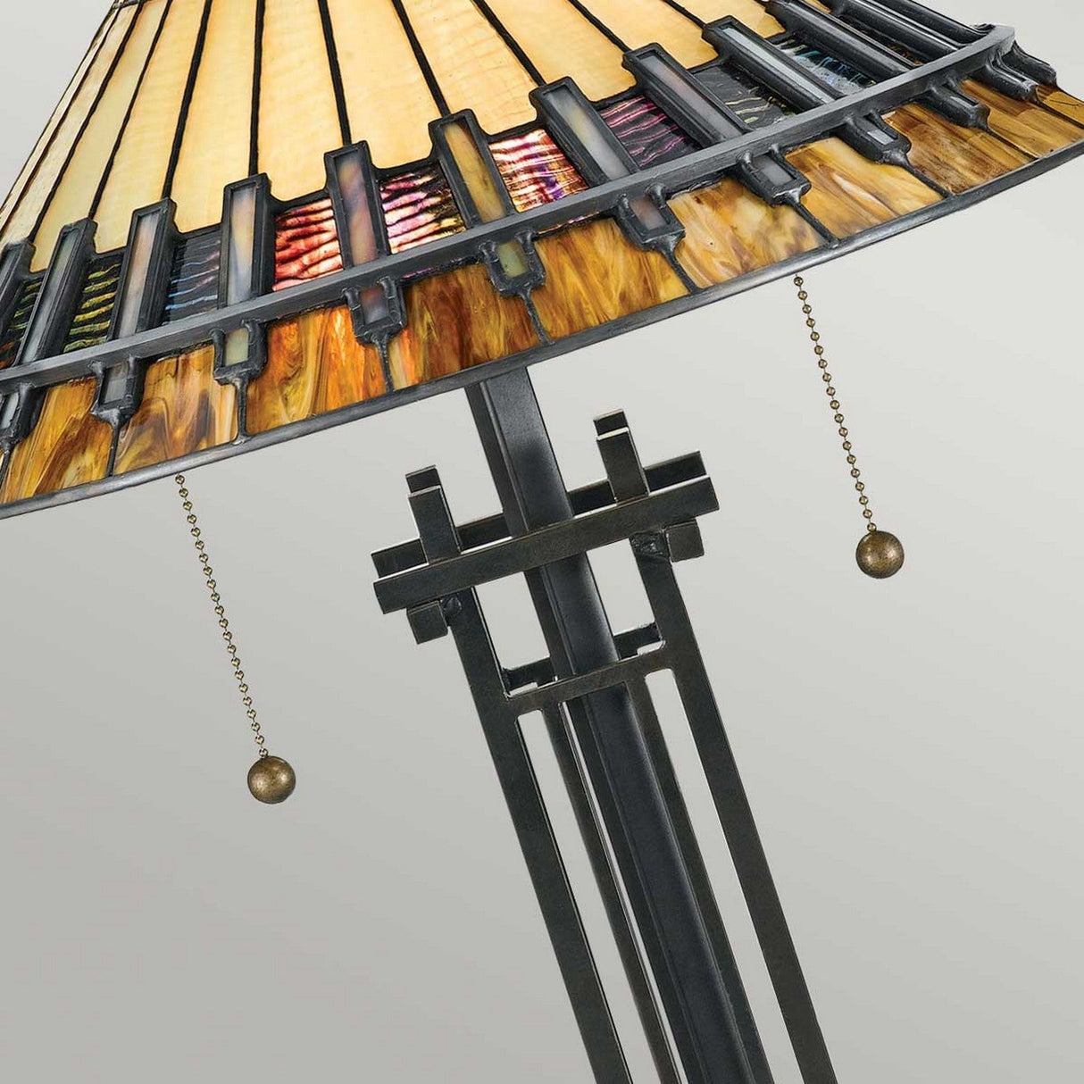 A Chastain 2 Light Tiffany Table Lamp in Vintage Bronze presents a decorative shade showcasing a geometric pattern in a variety of colors. It features two pull chains for switches and stands out beautifully against a plain, light background, harmonizing perfectly with its Arts and Crafts design.