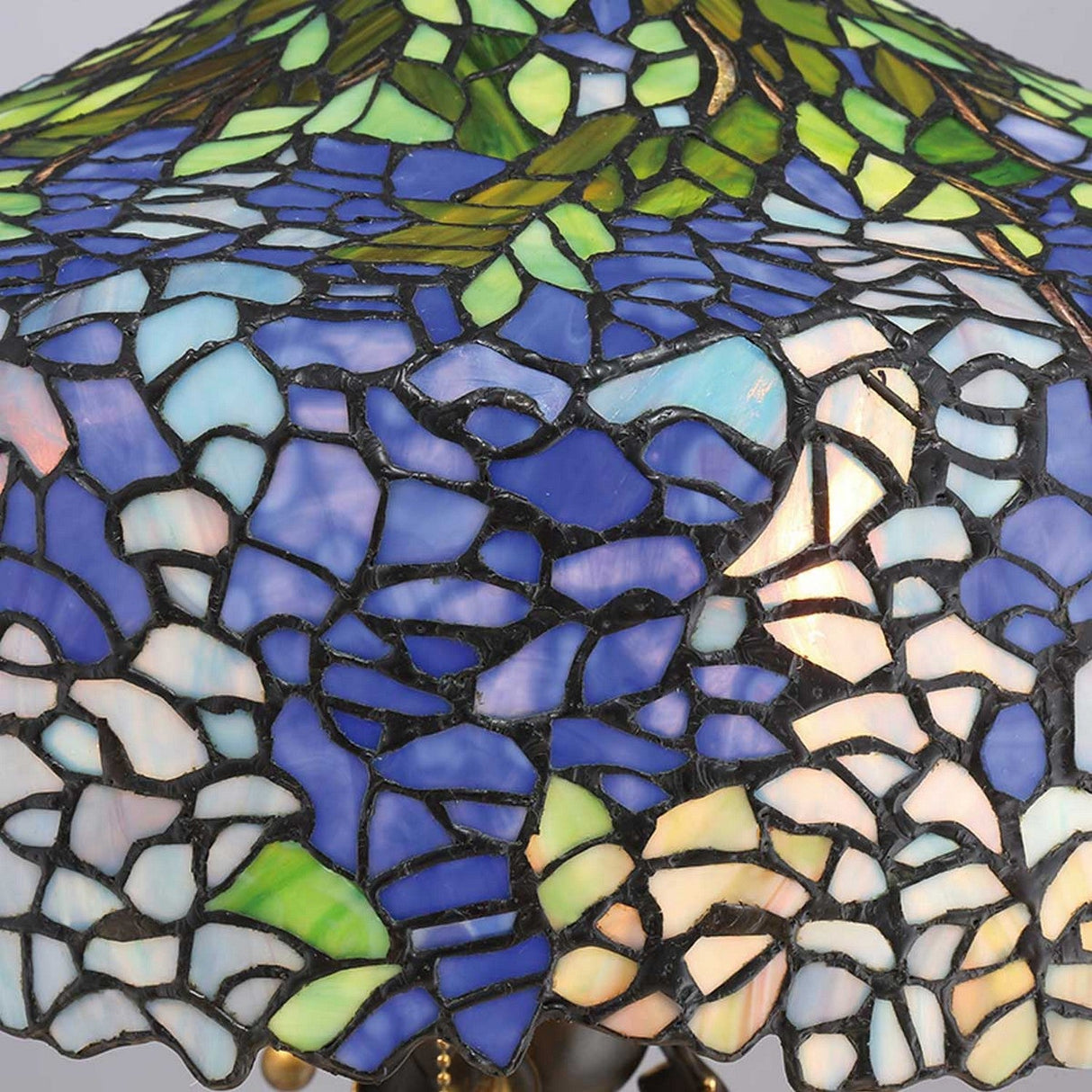 The Cobalt 3 Light Tiffany Table Lamp, with its vintage bronze finish, showcases a striking stained glass lampshade adorned in a mosaic pattern. It features blue, purple, green, and white pieces that come together to create an intricate design inspired by hydrangeas for a vibrant and colorful look.