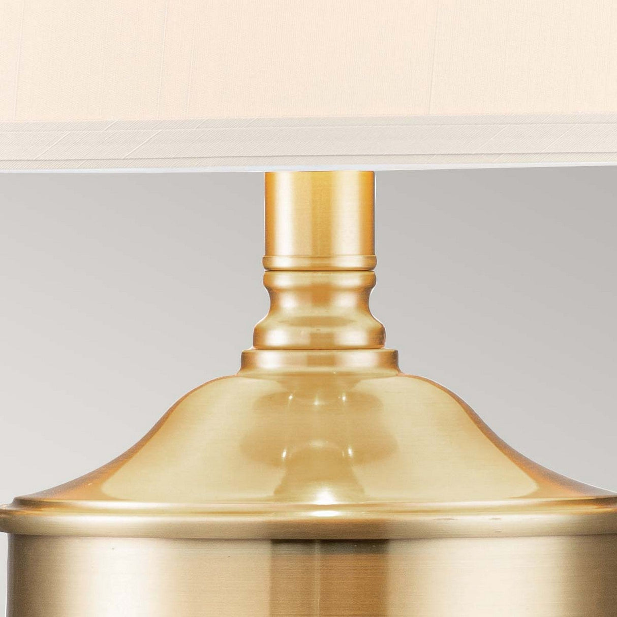 Close-up of the Dennison 1 Light Table Lamp - Brushed Brass, showcasing its smooth satin finish. The design includes a rounded top and subtle neck detailing, supporting a light-colored fabric lampshade. Ideal for home decor, it is set against a simple, neutral gray backdrop.