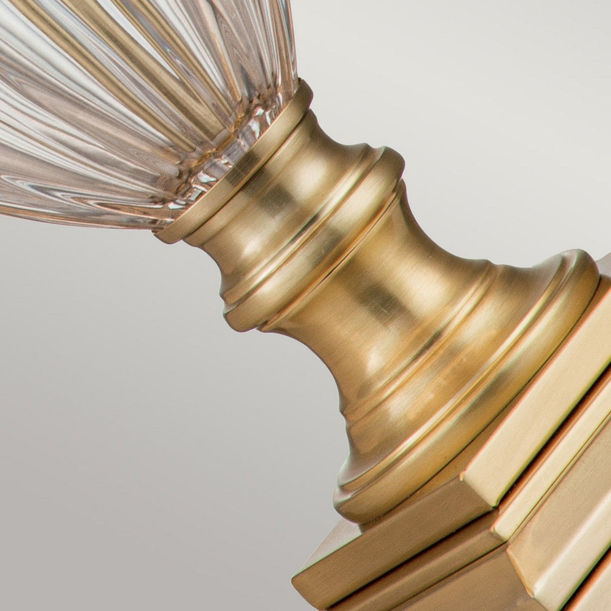 Close-up of the Dennison 1 Light Table Lamp in brushed brass, showcasing its detailed polished metalwork and ribbed glass stem. The lamp's textured, transparent glass enhances the sophistication of any home decor. The background features a soft gradient.