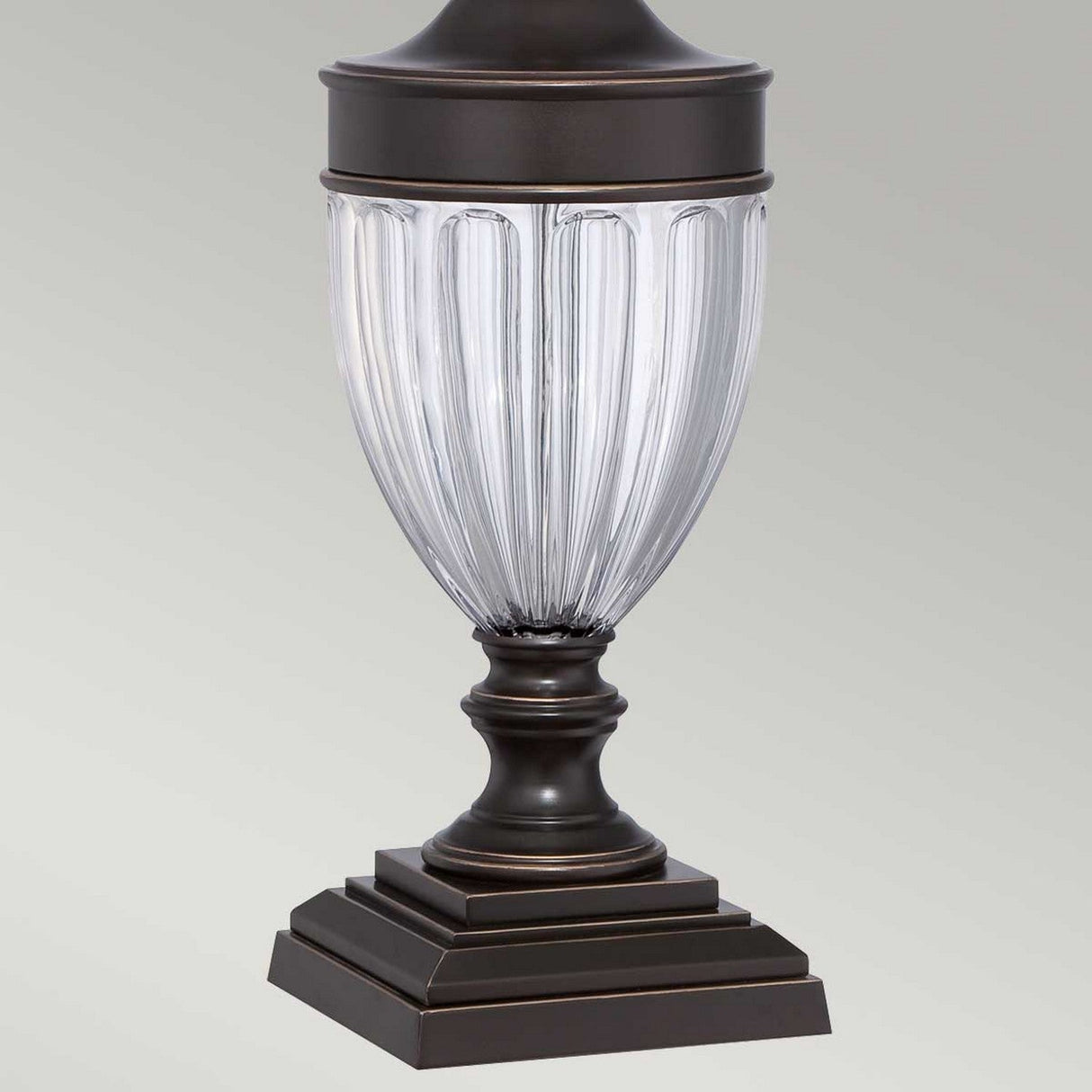 The Dennison 1 Light Table Lamp in Palladian Bronze boasts a decorative fluted design, making it ideal for transitional interiors. Its tiered square base and curved glass body are complemented by a luxurious Palladian bronze top, all set against a plain gray background that enhances its sophisticated charm.