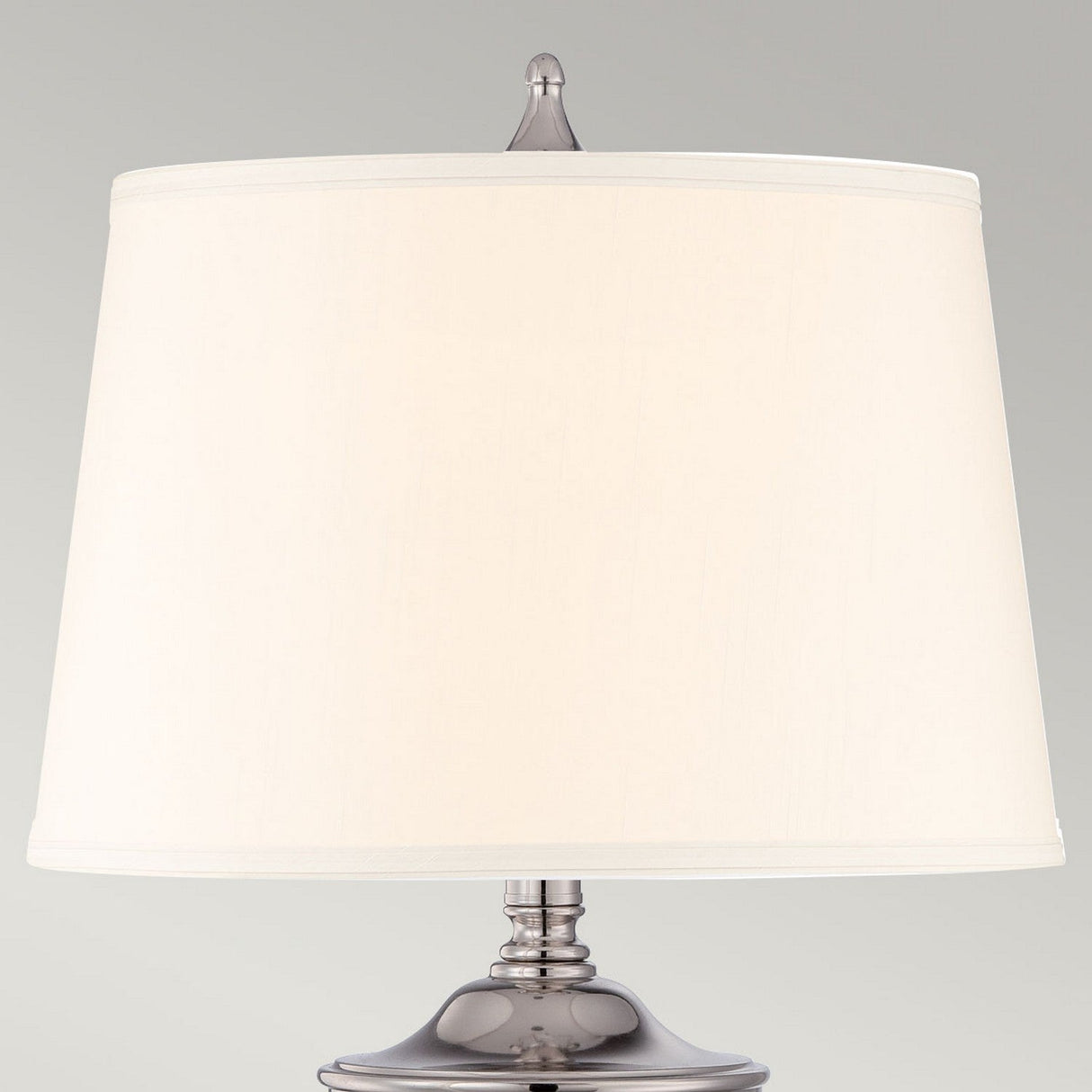A detailed view of the Dennison 1 Light Table Lamp - Polished Nickel reveals its modern design, featuring a white lampshade coupled with a metallic base finished in polished nickel. The minimalist light grey background enhances the ambient glow, underscoring the lamp's simplicity and elegance.
