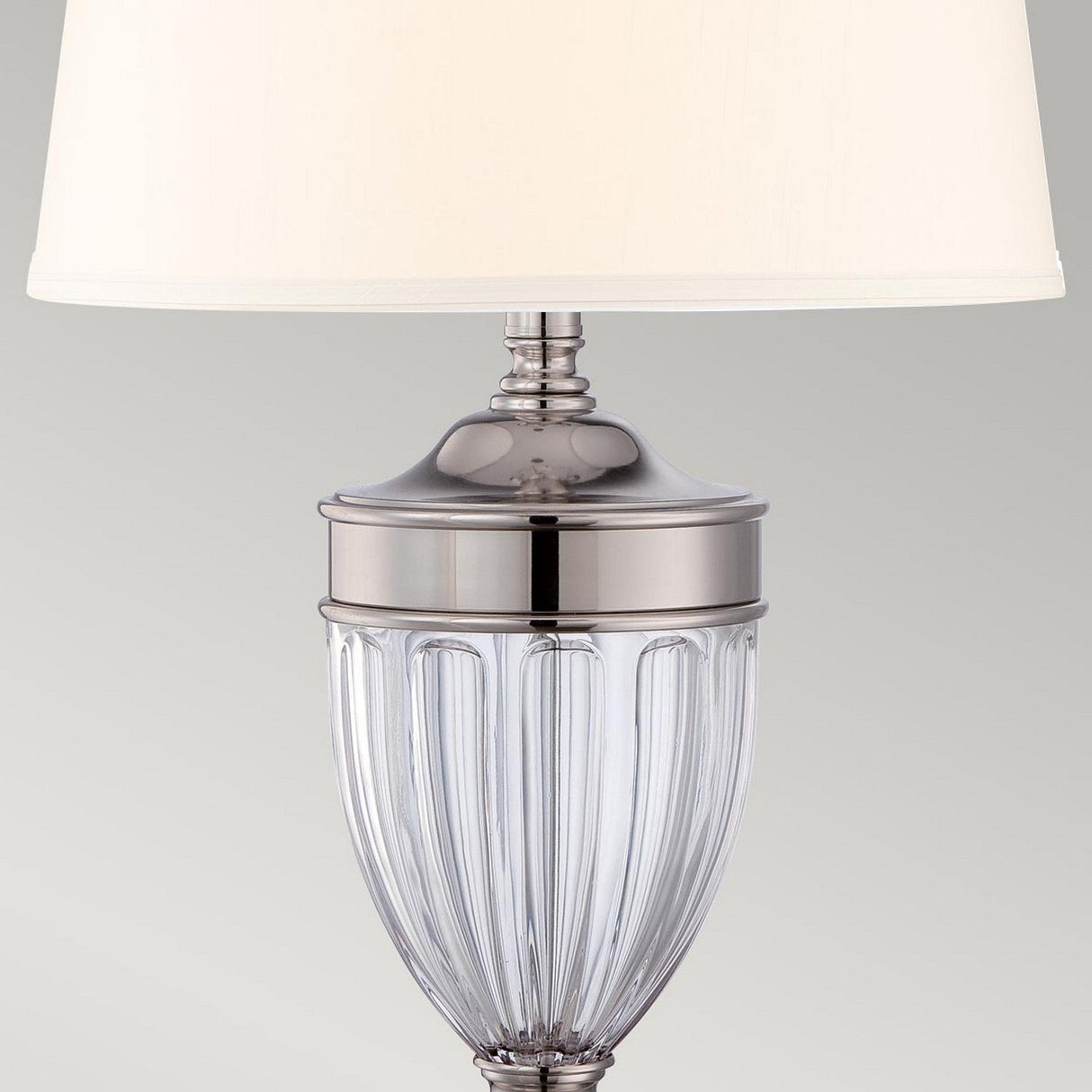The Dennison 1 Light Table Lamp - Polished Nickel features a glass base with vertical ribbed detailing and is complemented by a white lampshade. Its polished nickel finish adds a touch of elegance to its modern design, offering an ambient glow that suits any space.