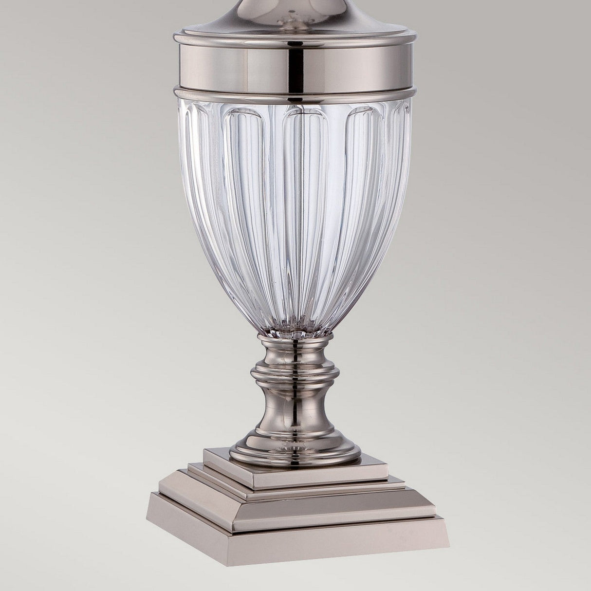 The Dennison 1 Light Table Lamp - Polished Nickel showcases a timeless metal design with an ornate clear glass body and decorative fluted details. It stands gracefully on a tiered square base, casting an ambient glow against a simple gray background.