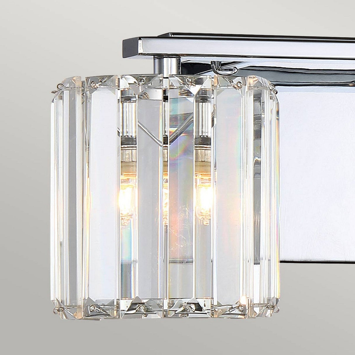 The Divine 2 Light Wall Light features vertical crystal glass slats surrounding two bulbs, casting a soft glow. Its sleek, polished chrome finish enhances its elegance against a plain background.