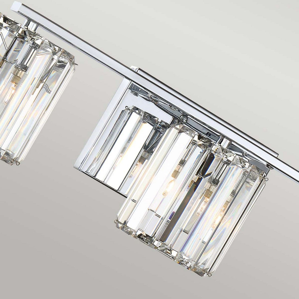 A detailed close-up of the Divine 3 Light Wall Light - Polished Chrome is depicted, highlighting its two cylindrical ribbed glass shades with exposed bulbs. The shades are elegantly affixed to a smooth horizontal metal bar, set against a simple backdrop.