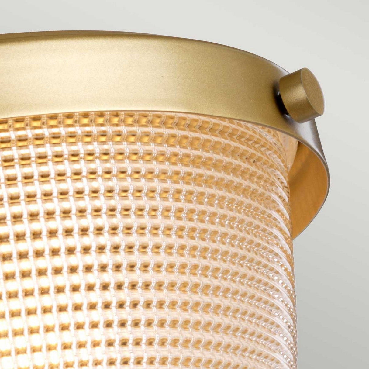This Dublin 1 Light Semi Flush - Natural Brass showcases a textured cylindrical glass design with a golden metallic rim, offering the look of natural brass. The grid pattern enhances its warm and slightly diffused glow, making it perfect against a light gray background for bathroom lighting.