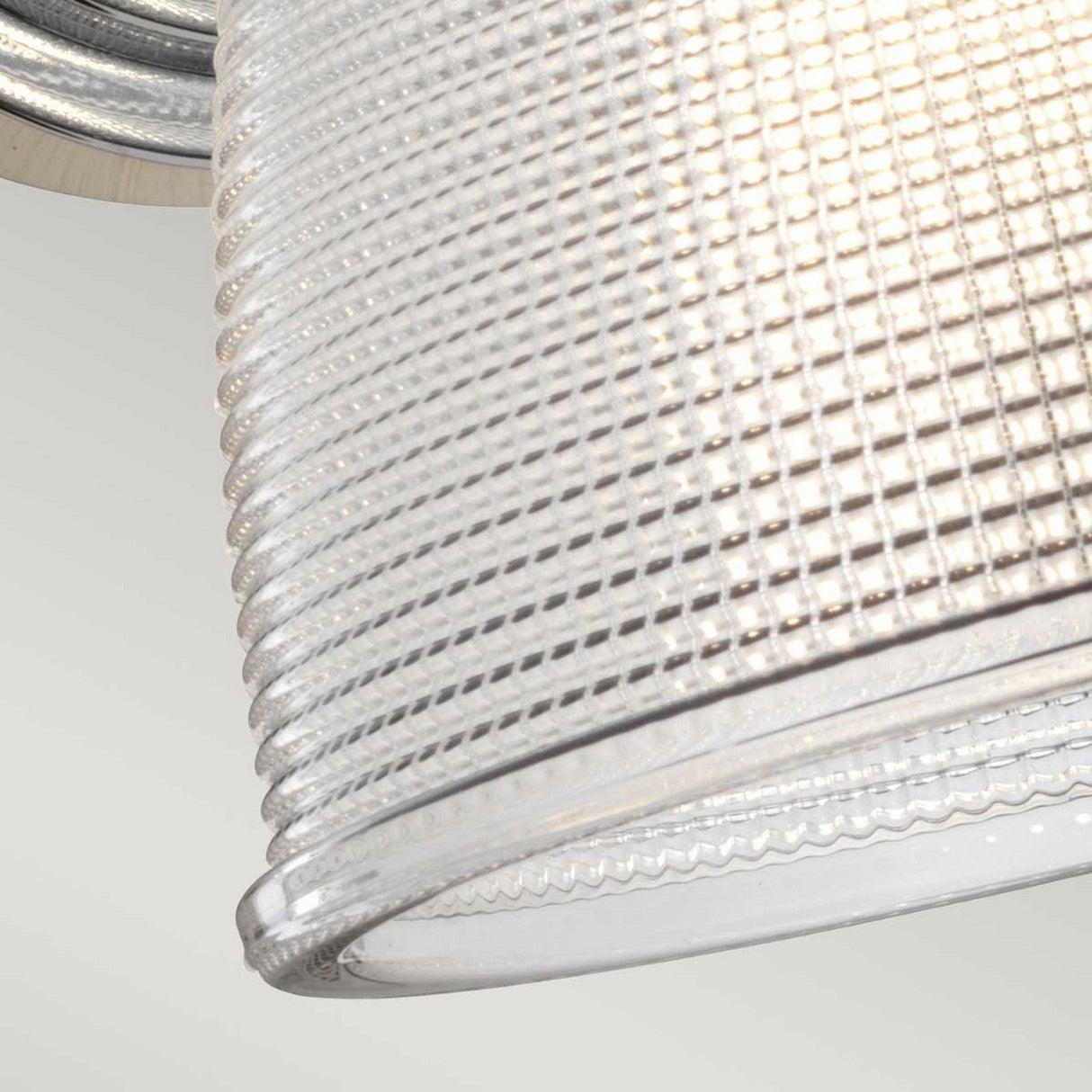 A close-up of the Dublin 1 Light Wall Light - Polished Chrome features a textured glass lampshade with a grid pattern, ideal for bathroom lighting. The illuminated lamp emits a warm glow through its translucent surface, with polished chrome accents enhancing the smooth, rounded edge of the shade.