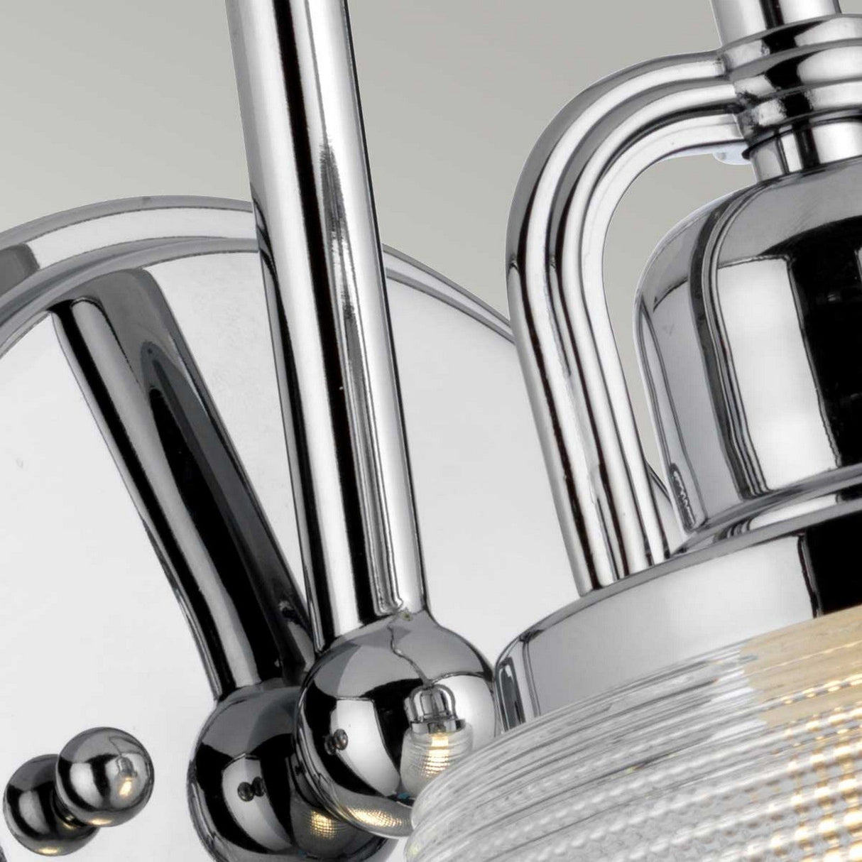 Close-up of the Dublin 1 Light Wall Light in polished chrome, showcasing its ribbed glass shade. The wall light's reflective surface and sleek metal design offer an elegant contrast against the soft gray background, making it an ideal choice for vanity lighting.