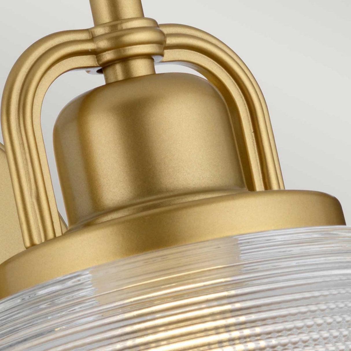 A close-up of the Dublin 1 Light Wall Light showcases its gold metallic finish, with a ribbed glass lampshade. The design includes curved supports and exudes a modern, industrial style, making it ideal for bathroom lighting. Its natural brass surfaces reflect light beautifully, enhancing the texture and shine of the metal finish.