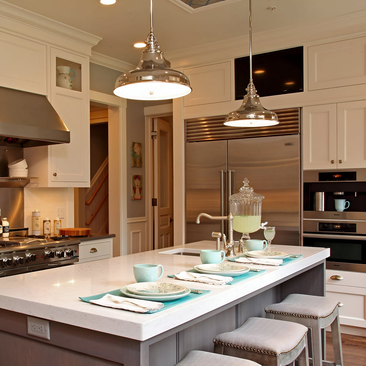A contemporary kitchen includes a spacious island topped with a white countertop, adorned with teal placemats, cups, and plates. The Emery 1 Light Medium Pendant in Imperial Silver overhead adds sophistication. Stainless steel appliances, such as a fridge, oven, and stove, are arranged along the walls.